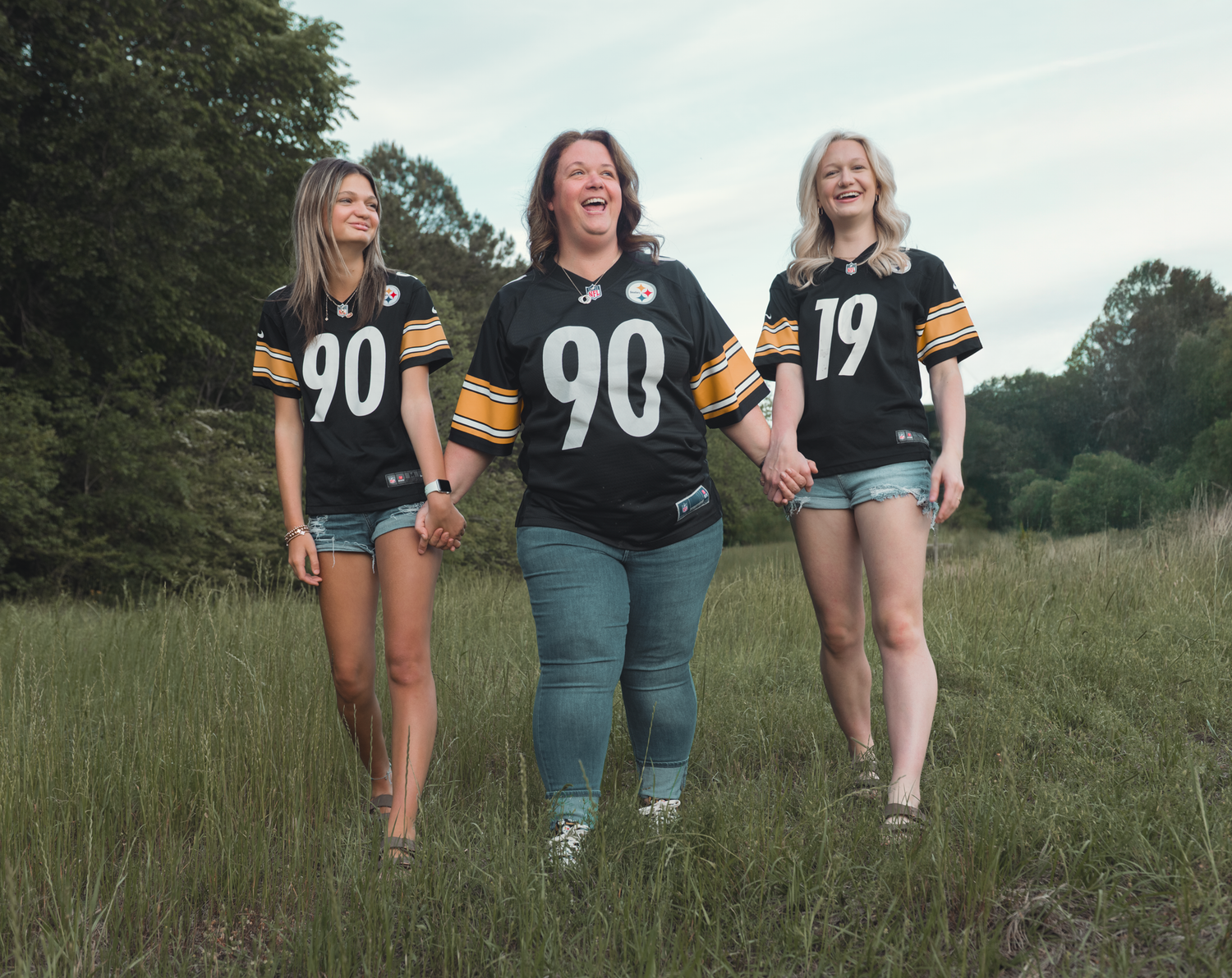 Three sports fans wearing black jerseys with number 90 and 19 pose together in a grassy field at dusk.