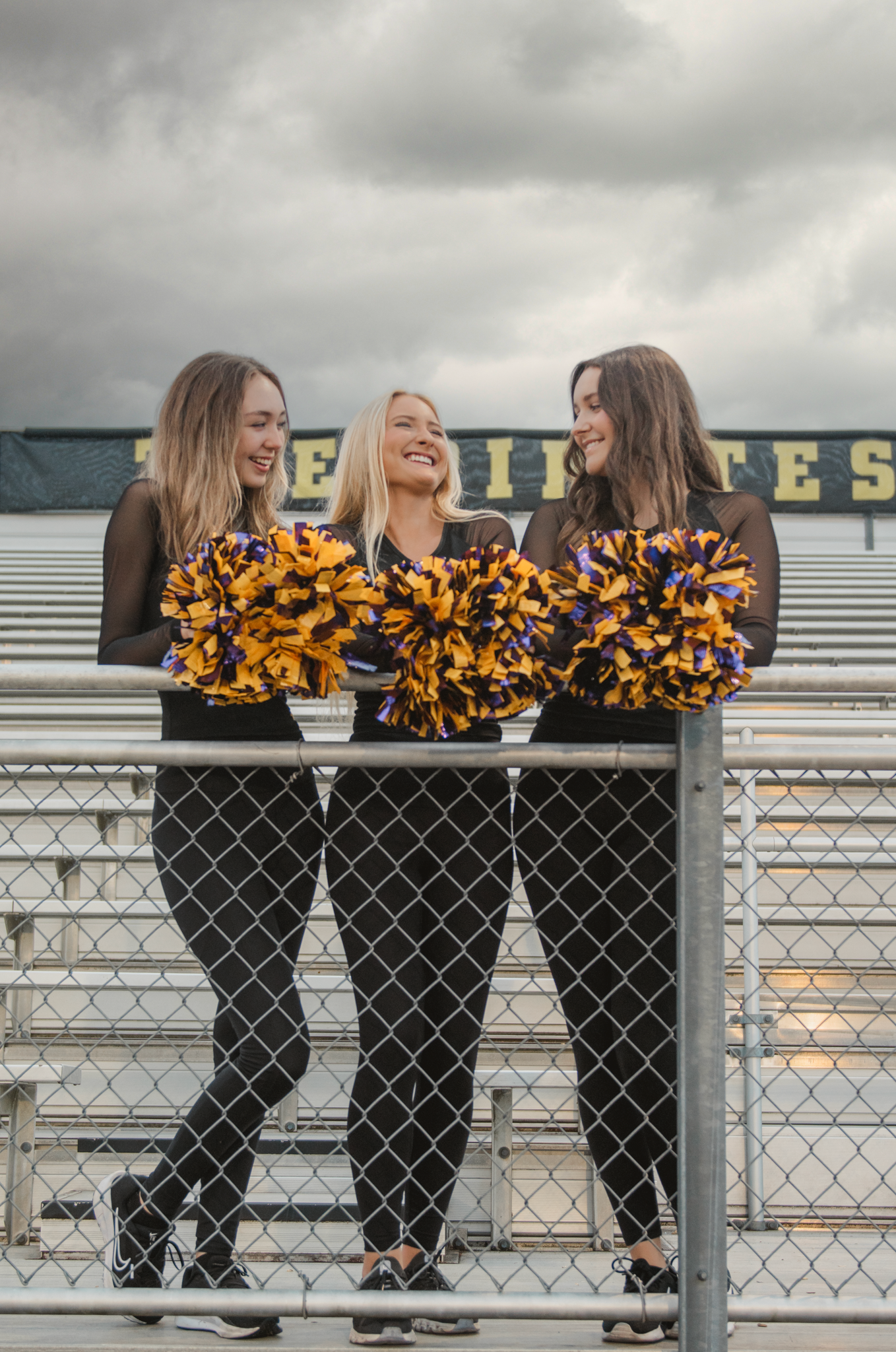 Cheerleaders in black outfits with gold pom-poms lean against a chain link fence while sharing a laugh together.