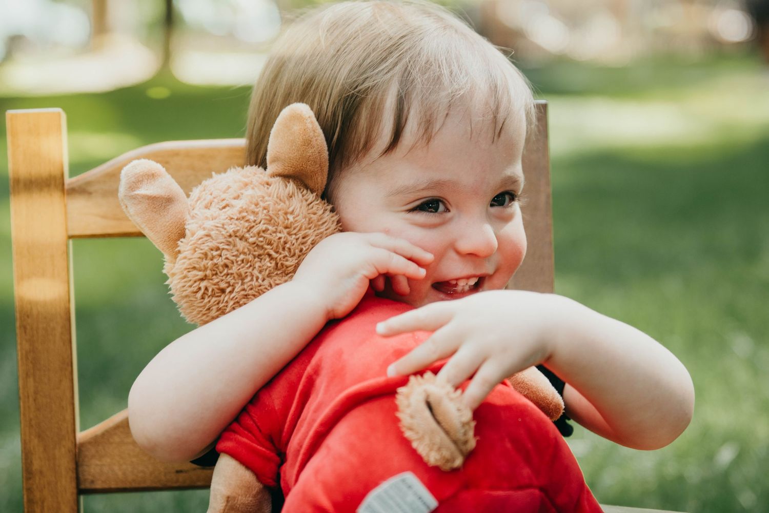A child in a red shirt sits in a wooden chair outdoors while hugging a teddy bear and smiling.