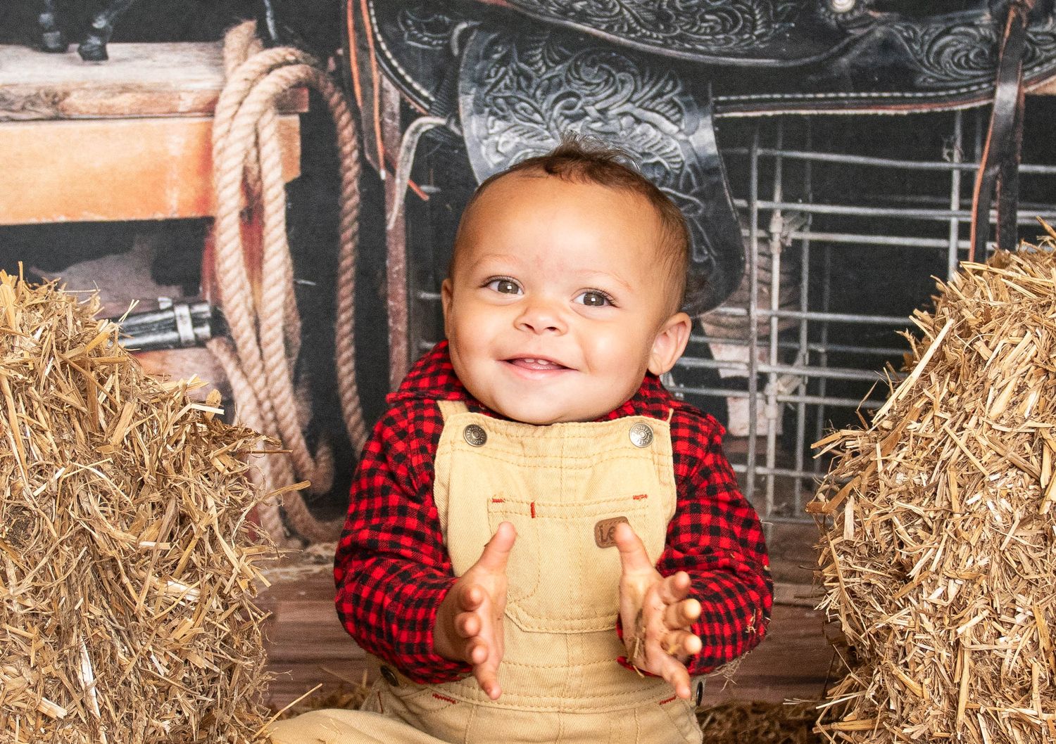 A young child in red buffalo plaid and tan overalls sits among hay bales in a rustic setting.