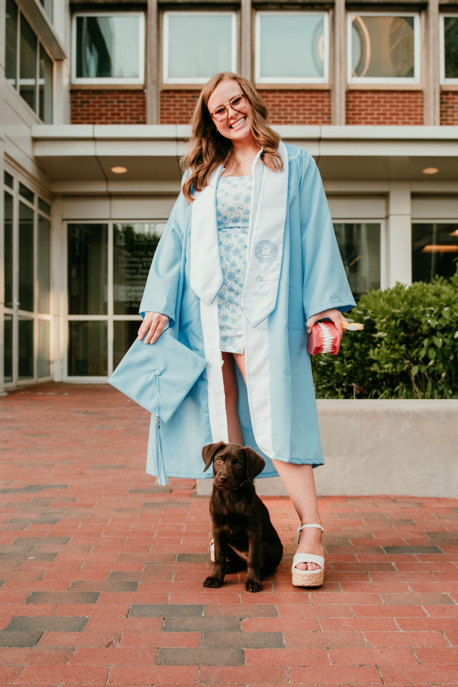 A graduation portrait with a chocolate lab puppy on red brick pavement, featuring light blue robes and casual sandals.