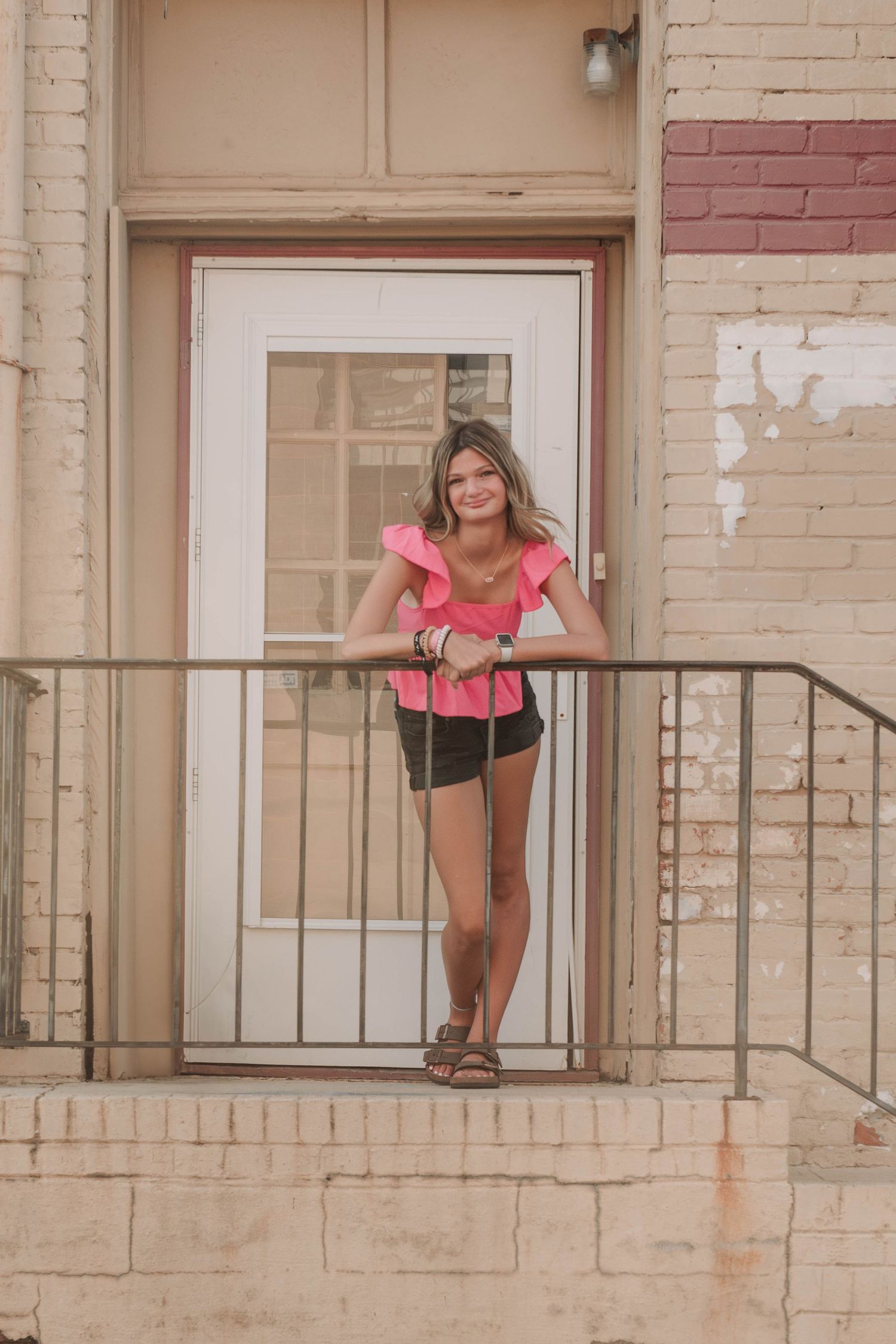 A person in a pink top and black shorts stands on an outdoor staircase landing against a beige brick building.