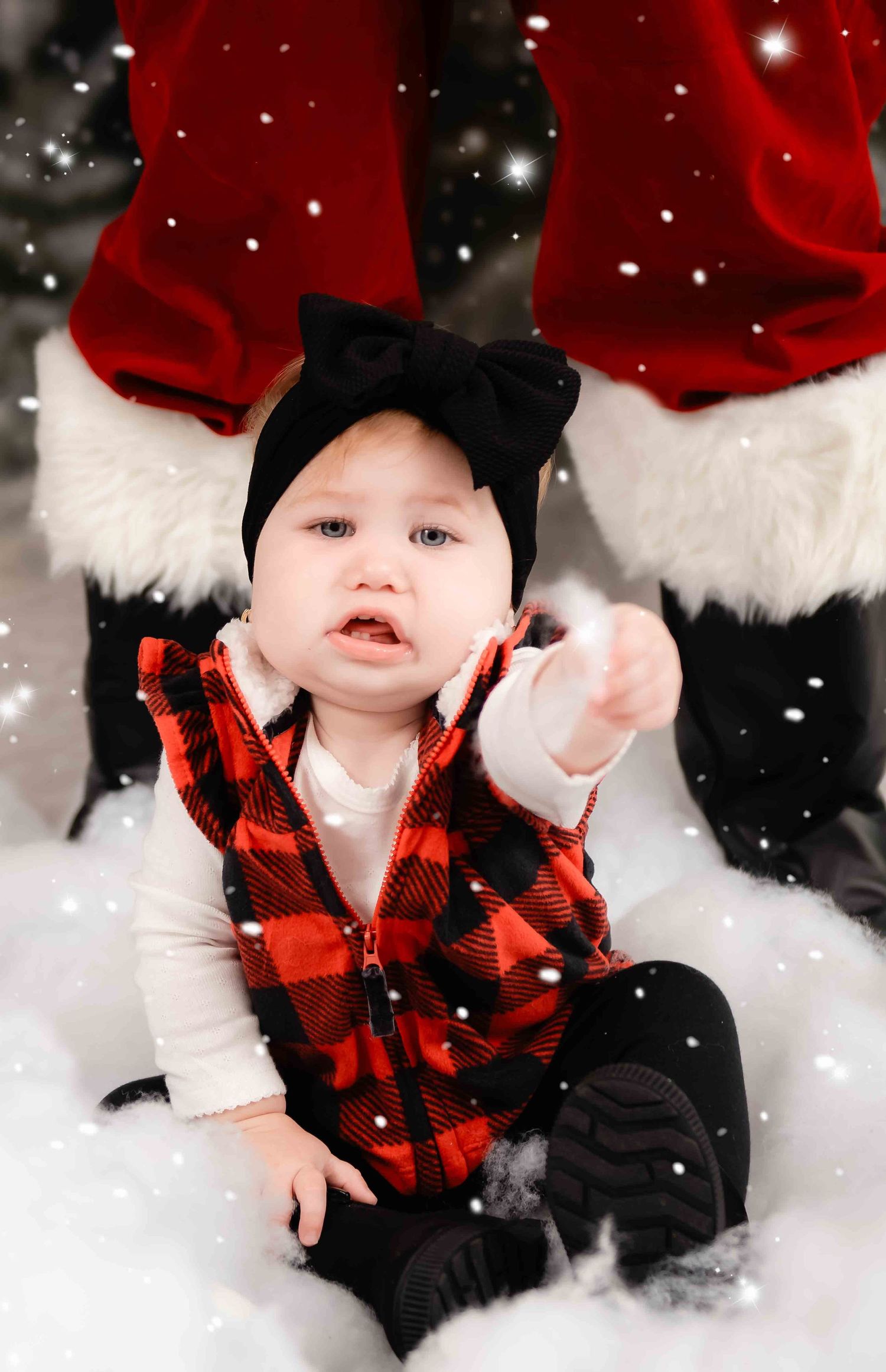 A young child in red plaid vest and black bow sits in snow with falling snowflakes during winter portraits.