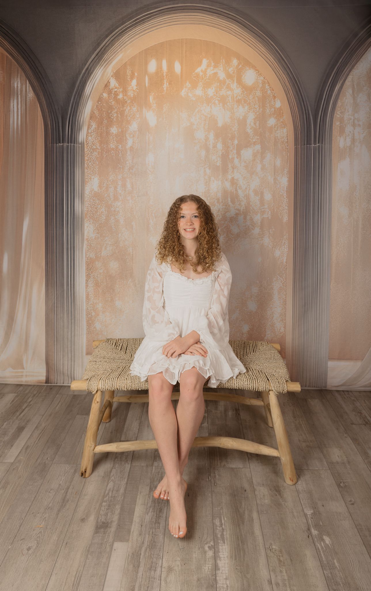 Figure in white dress sits barefoot on wooden bench against arched wall backdrop with textured beige surface.