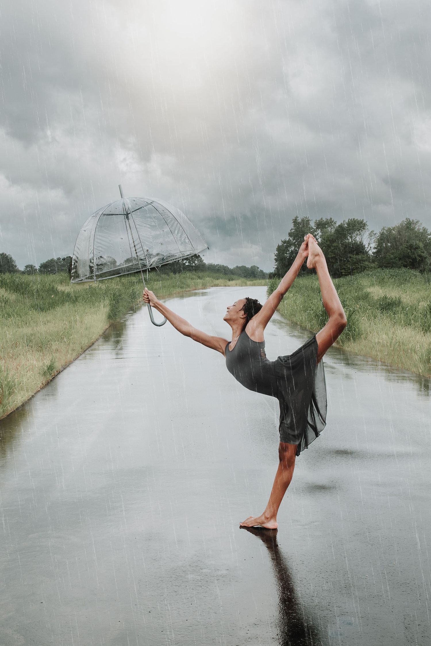 A person in black dances joyfully with a clear umbrella in the rain on a rural road.
