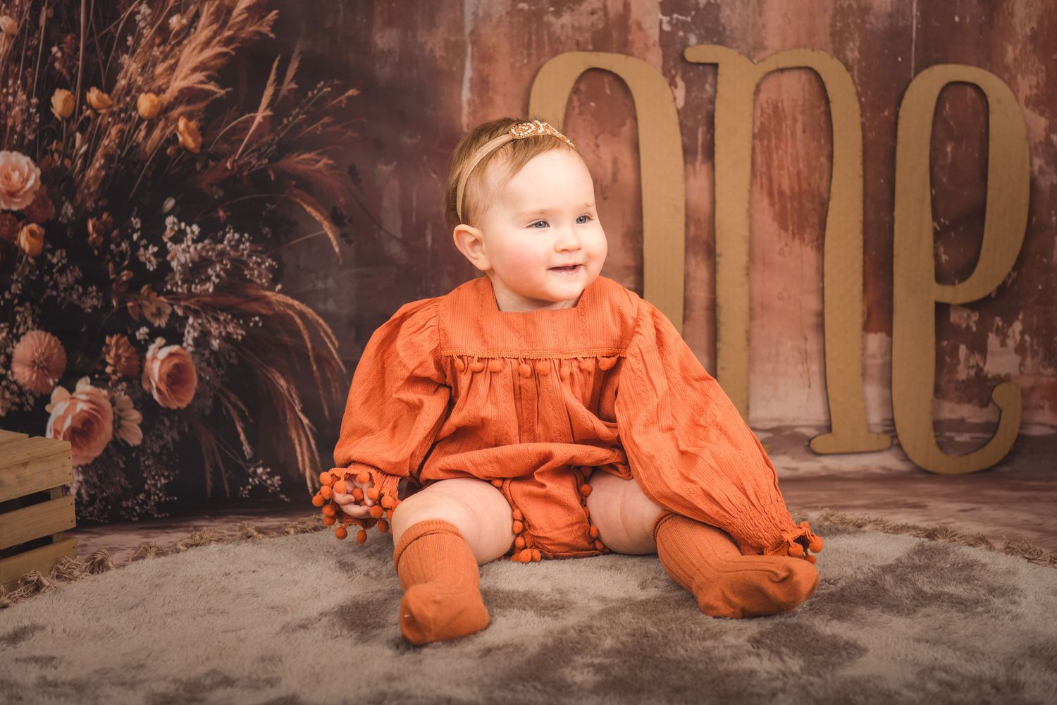 A small child wearing a bright orange bonnet and romper sits on a gray surface with floral decorations in the background.