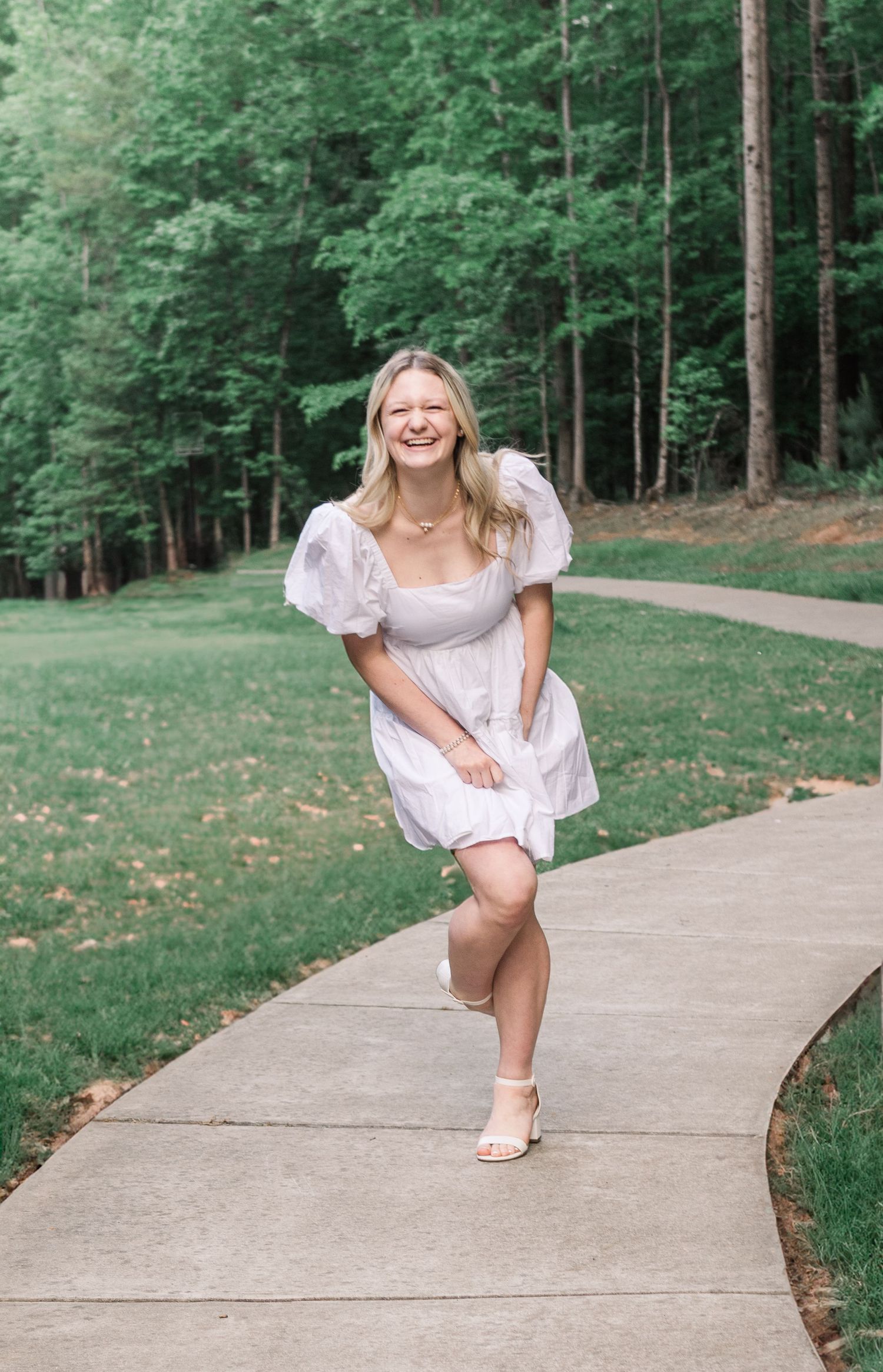 A person in a white summer dress poses playfully on a curved path surrounded by lush green forest.