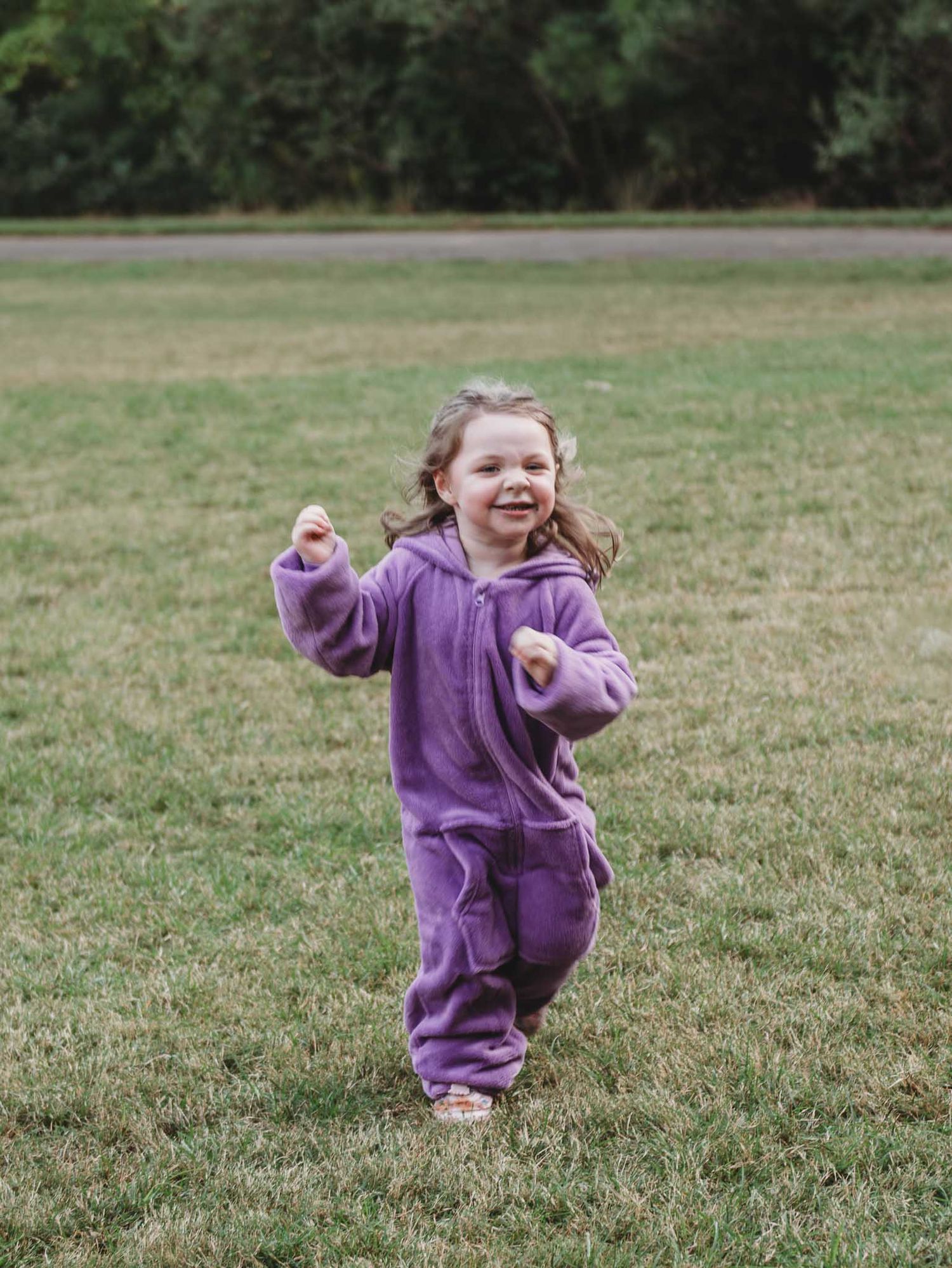 Energetic child runs and plays wearing a cozy purple fleece jumpsuit on a grassy field.