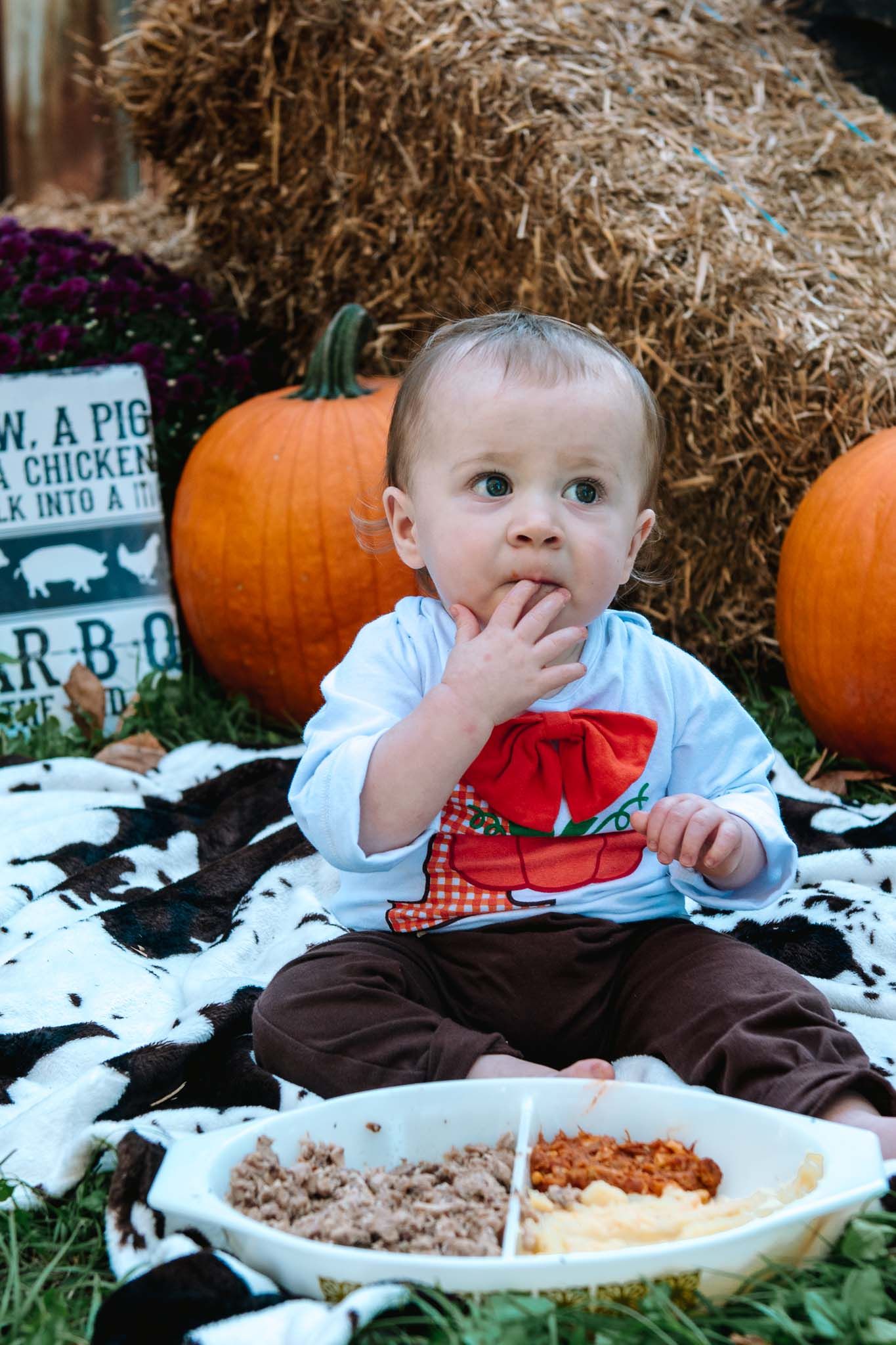 Autumn festive scene with pumpkins, hay bales, and a small child sitting on a blanket with snacks.