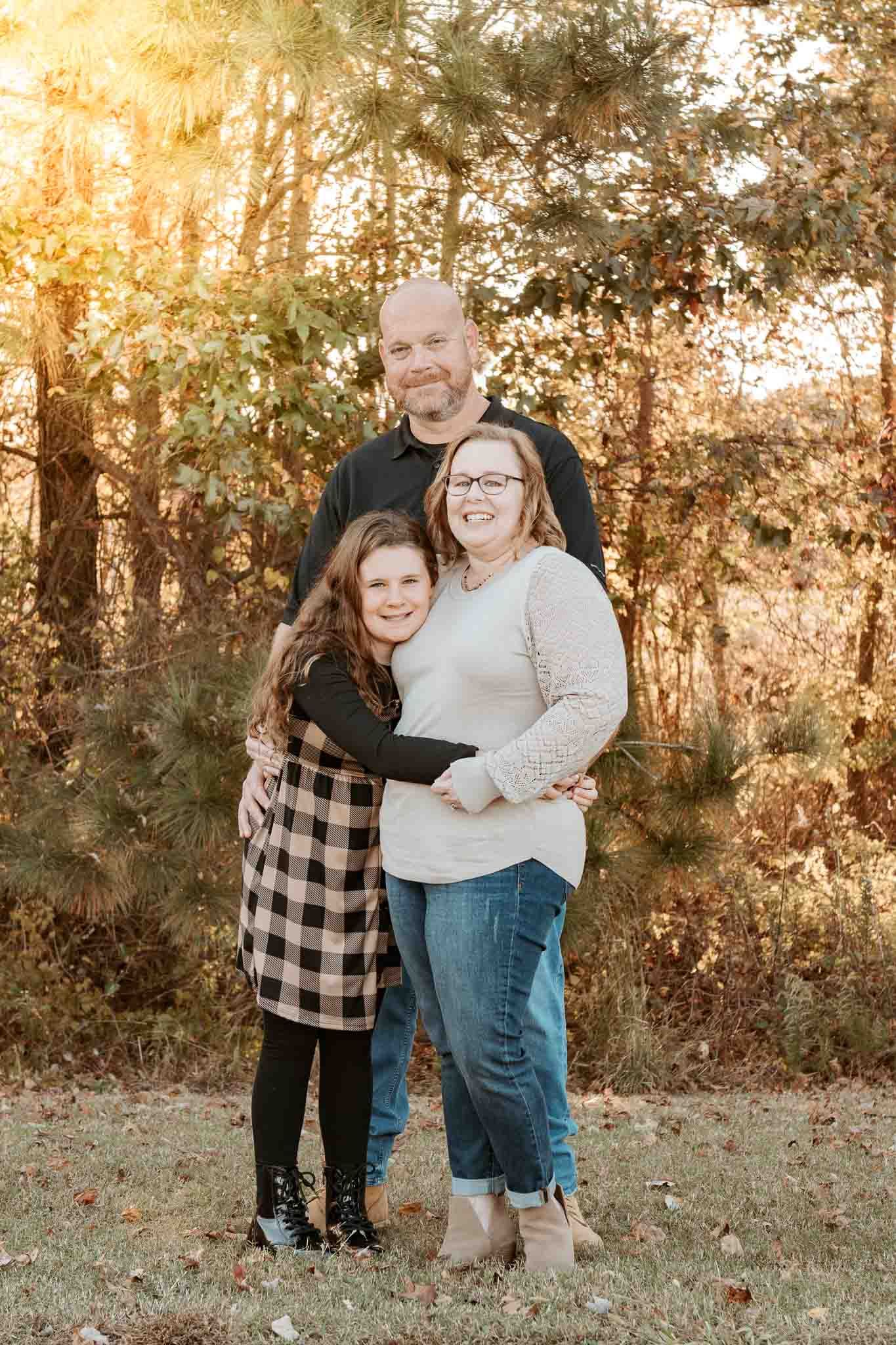 A family poses together in a wooded outdoor setting during golden hour with sunlight filtering through the trees.