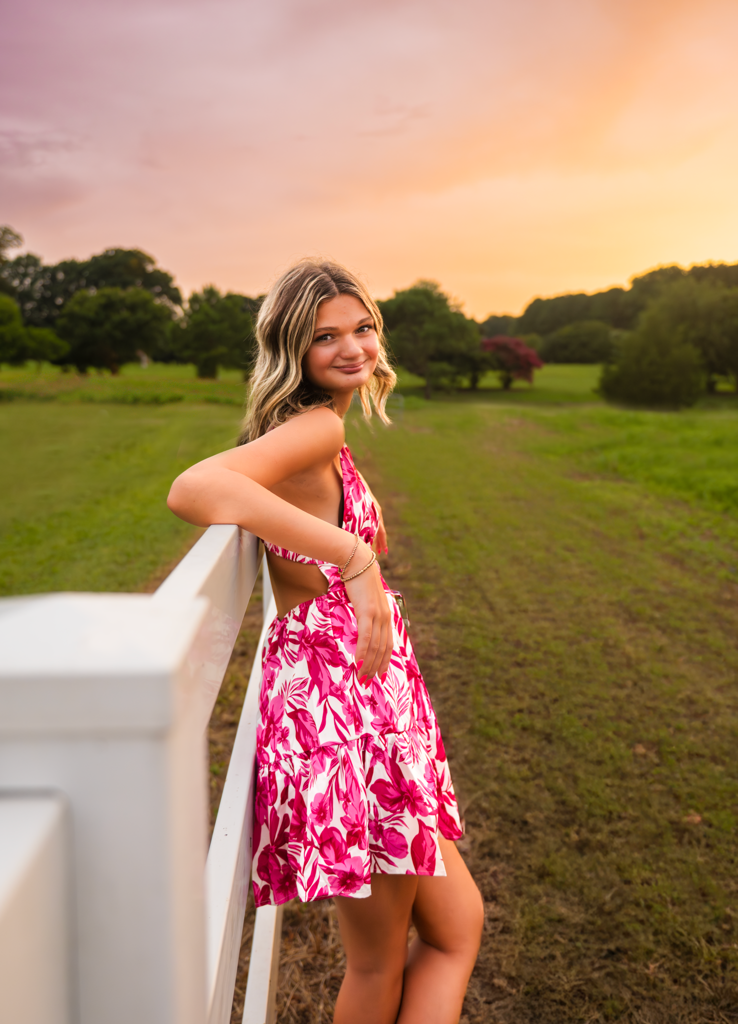 Person in a vibrant pink floral dress poses by a white fence at sunset in a grassy field.