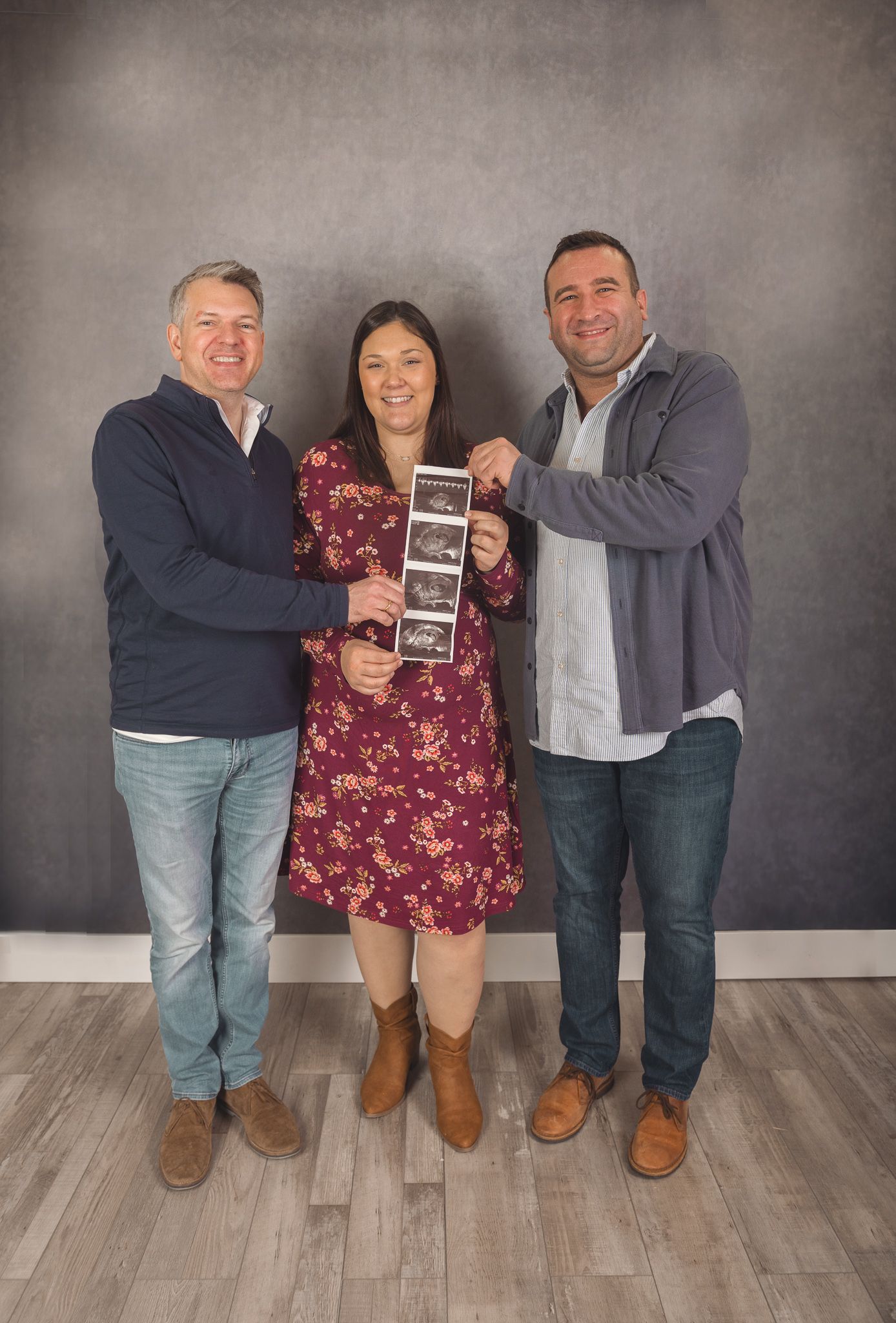 Three people stand together smiling against a gray backdrop while holding an ultrasound photo.