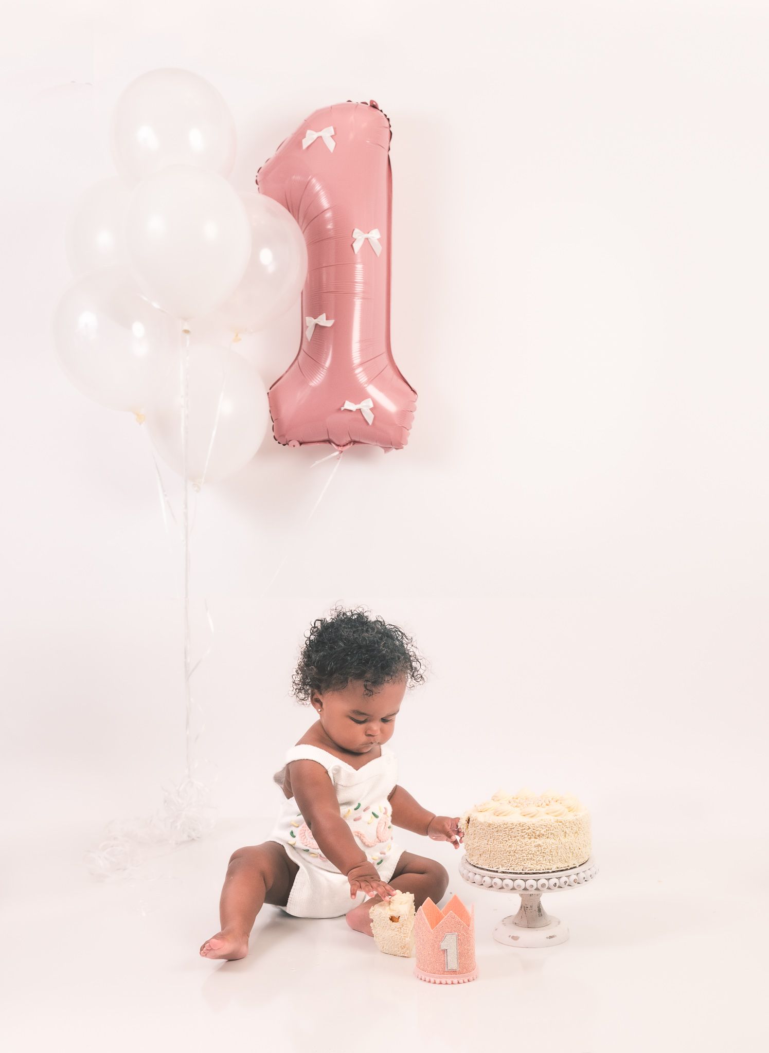 Baby in white outfit sitting on white backdrop with pink number one balloon and birthday cake for first cake smash photo.