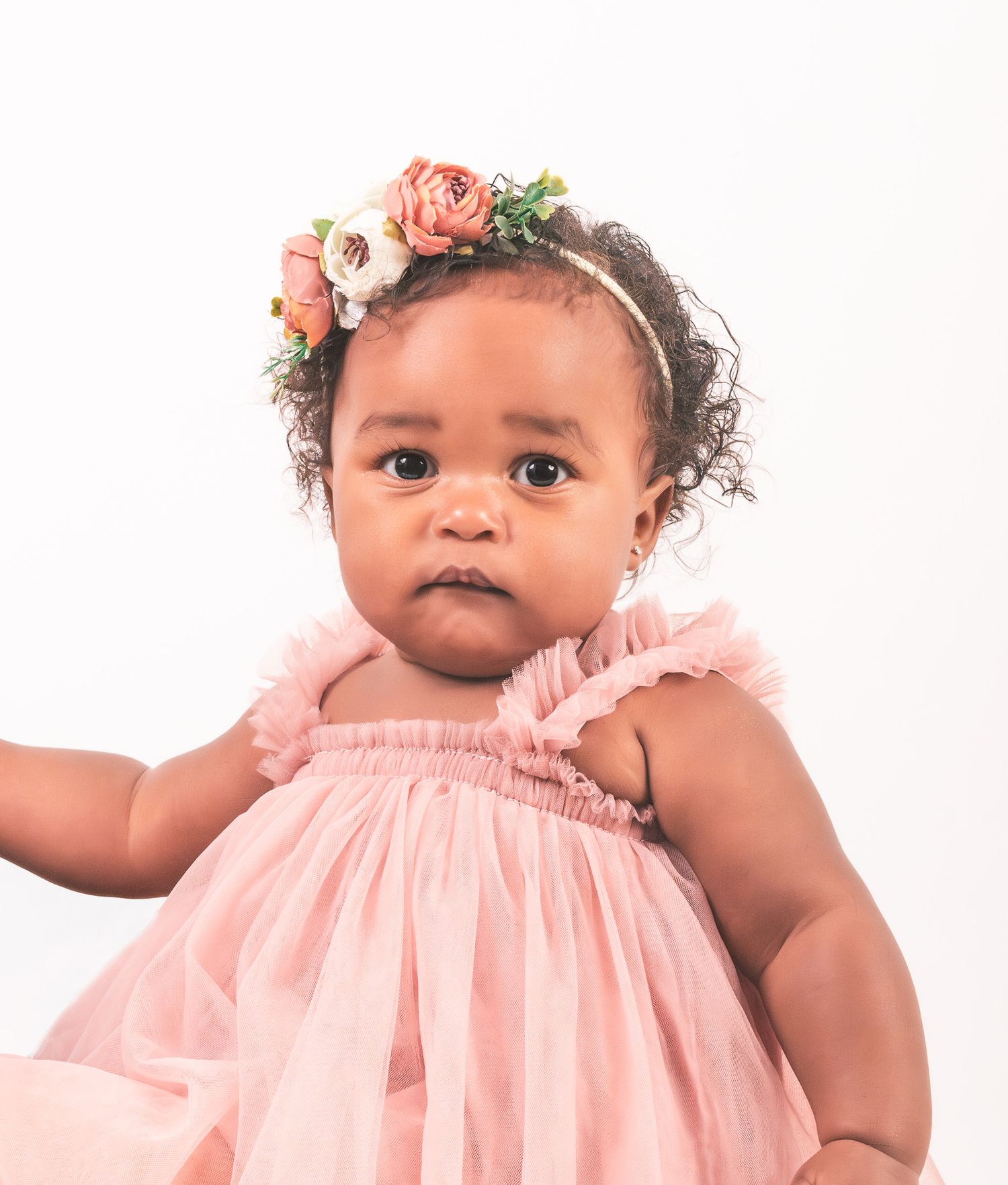 Baby wearing a pink ruffle dress and floral headband gazes at camera against white background.