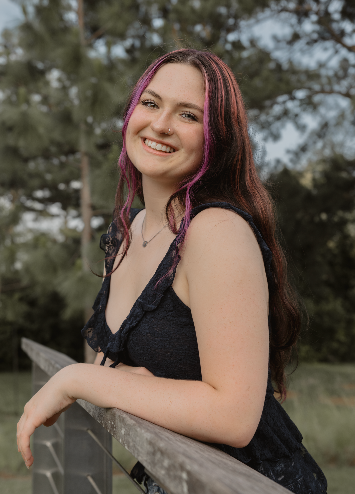 Haylee with purple-streaked hair smiles while leaning on wooden fence in an outdoor nature setting during her senior session.