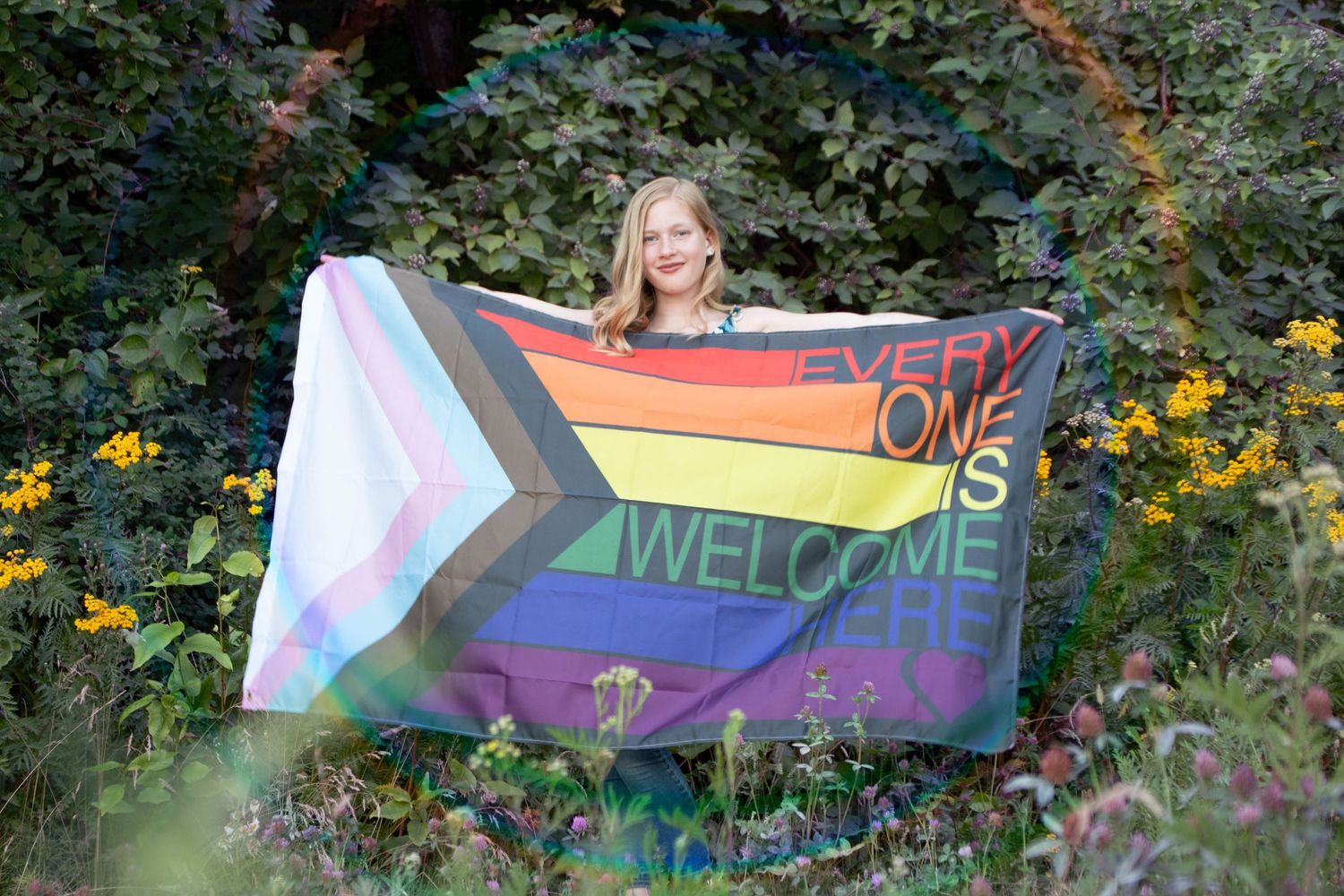 A colorful pride flag with 'WELCOME' text is displayed in a garden setting among yellow flowers and greenery.