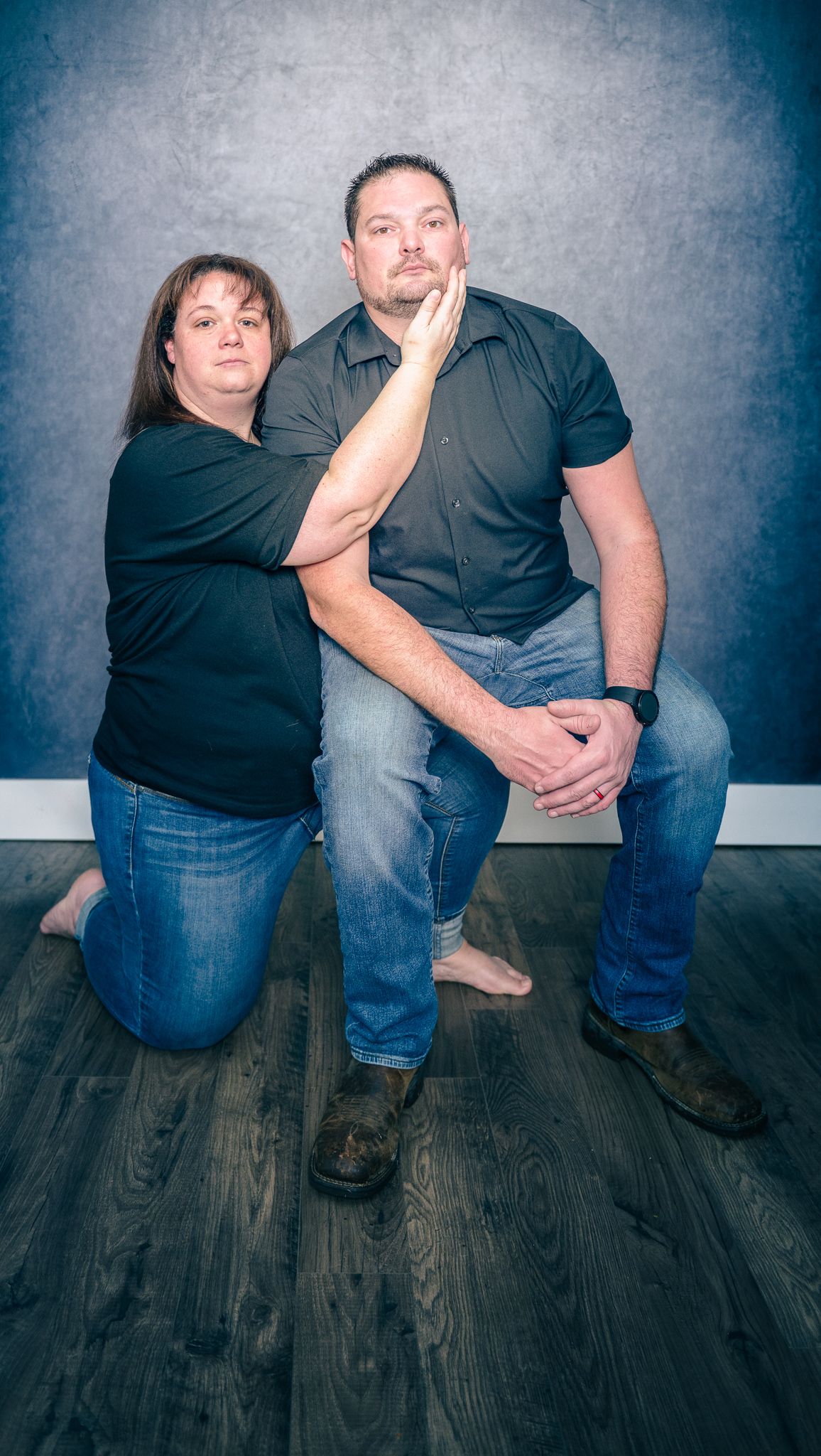 A couple poses together on a wooden floor against a gray studio background in casual attire.