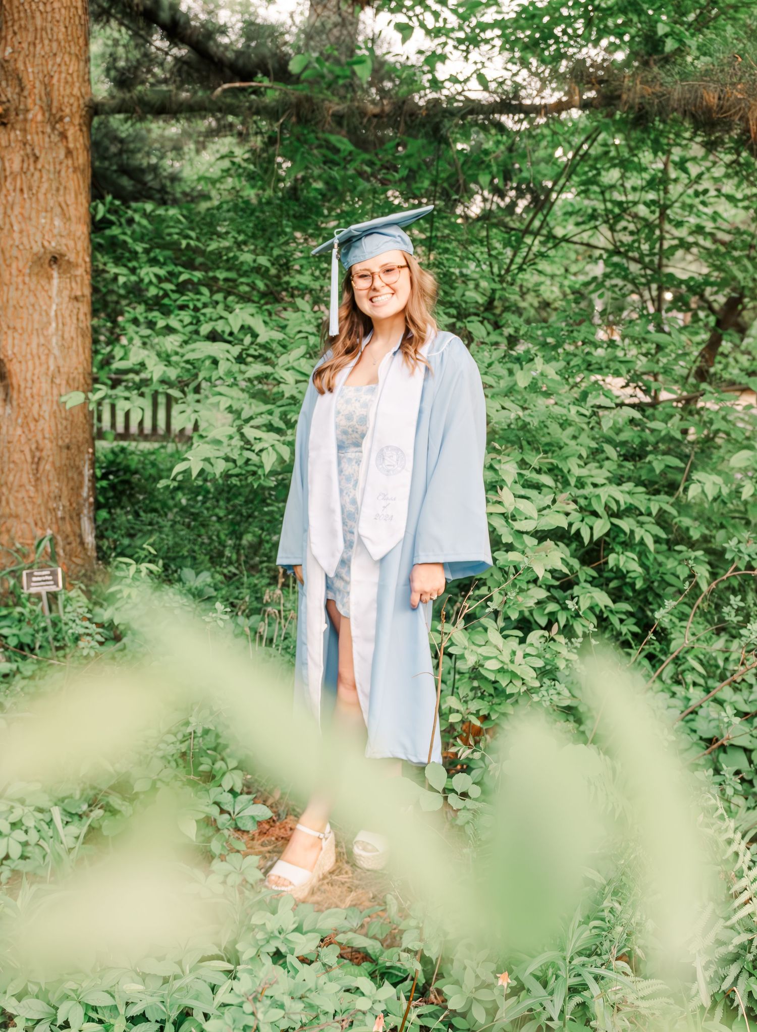 Graduate in light blue cap and gown poses in a lush green forest setting with natural sunlight filtering through trees.
