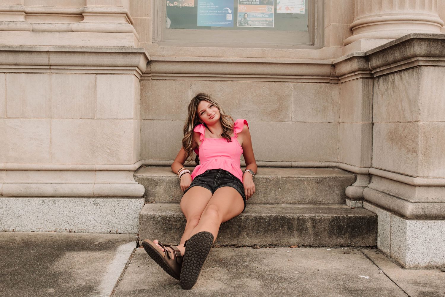 A figure relaxes on stone building steps wearing a pink sweater, black shorts and knee-high socks in an urban setting.
