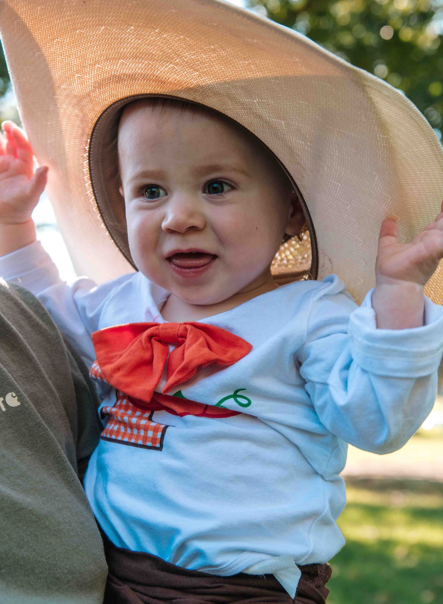 A baby in a floppy beige sun hat and light blue shirt with red bowtie smiles while being held outdoors.