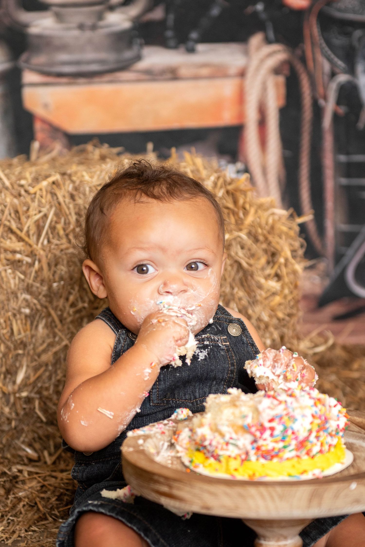 Small child in navy overalls enjoying a messy cake smash photo session while sitting on hay bales in rustic setting in Smithfield, NC