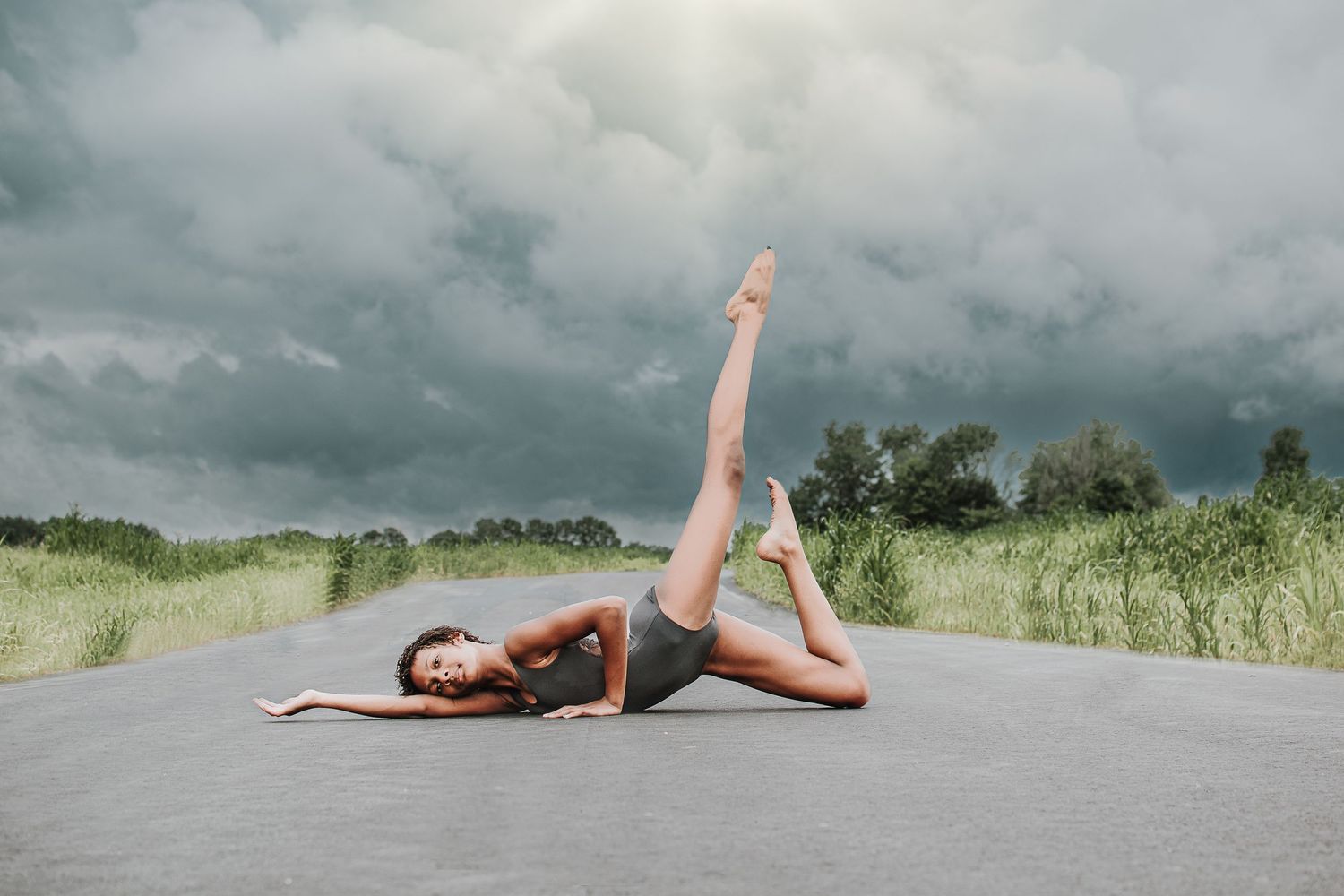 A dancer performs a leg lift pose outdoors on a concrete path beneath dramatic storm clouds.