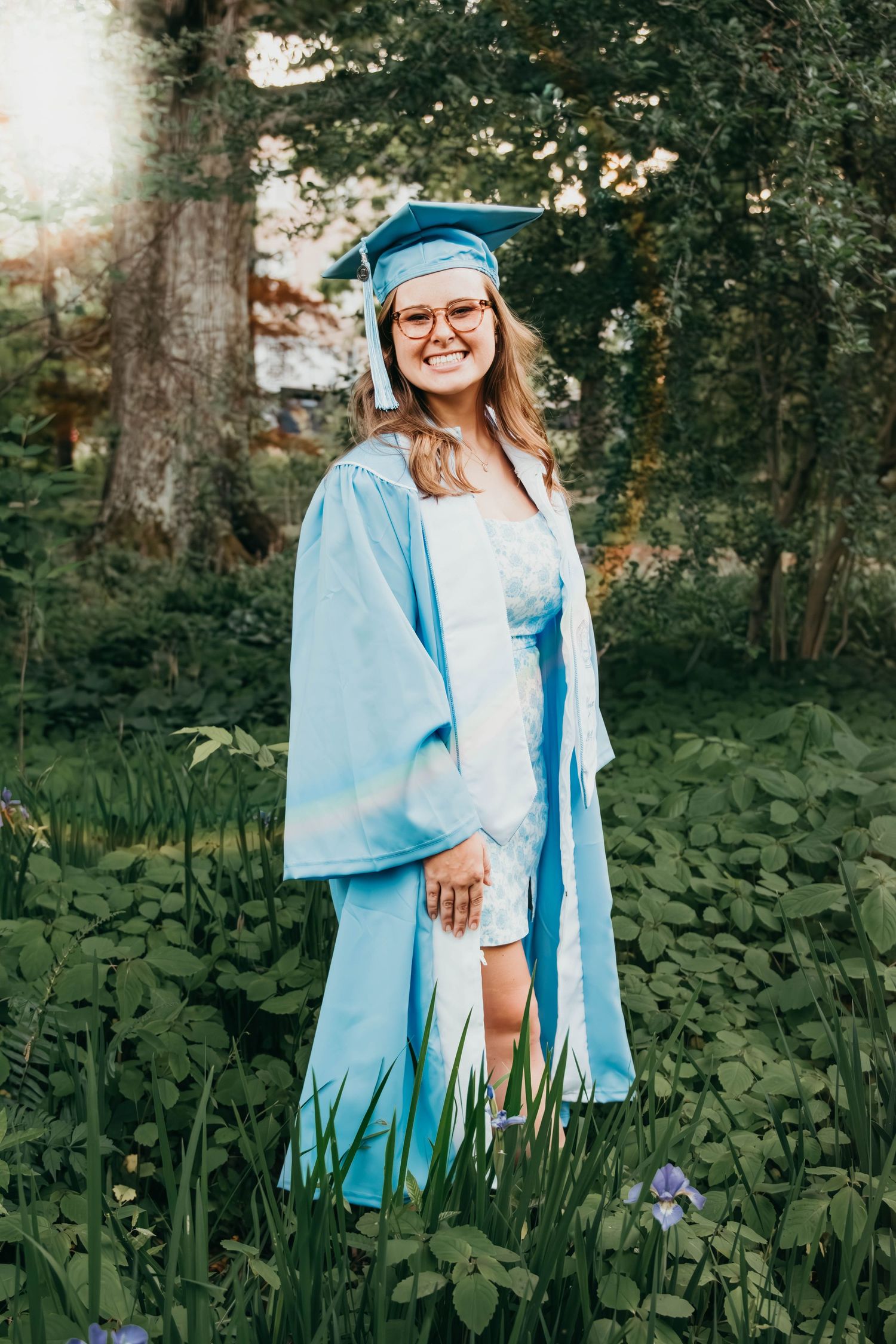 A graduate in light blue cap and gown stands outdoors in a lush garden setting during a sunny day.