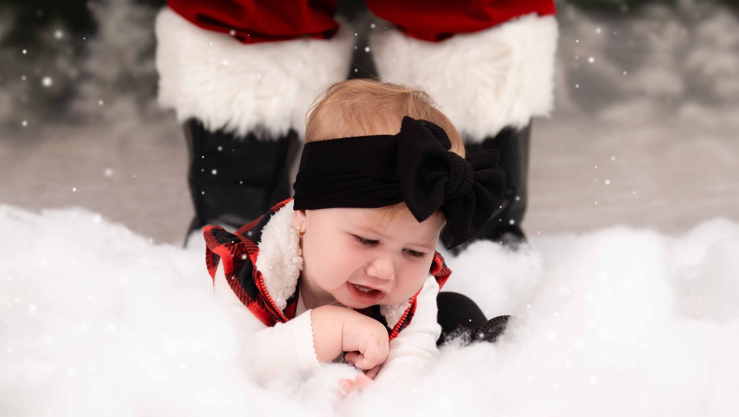 Adorable baby in black winter hat lying in snow with festive holiday decorations nearby.