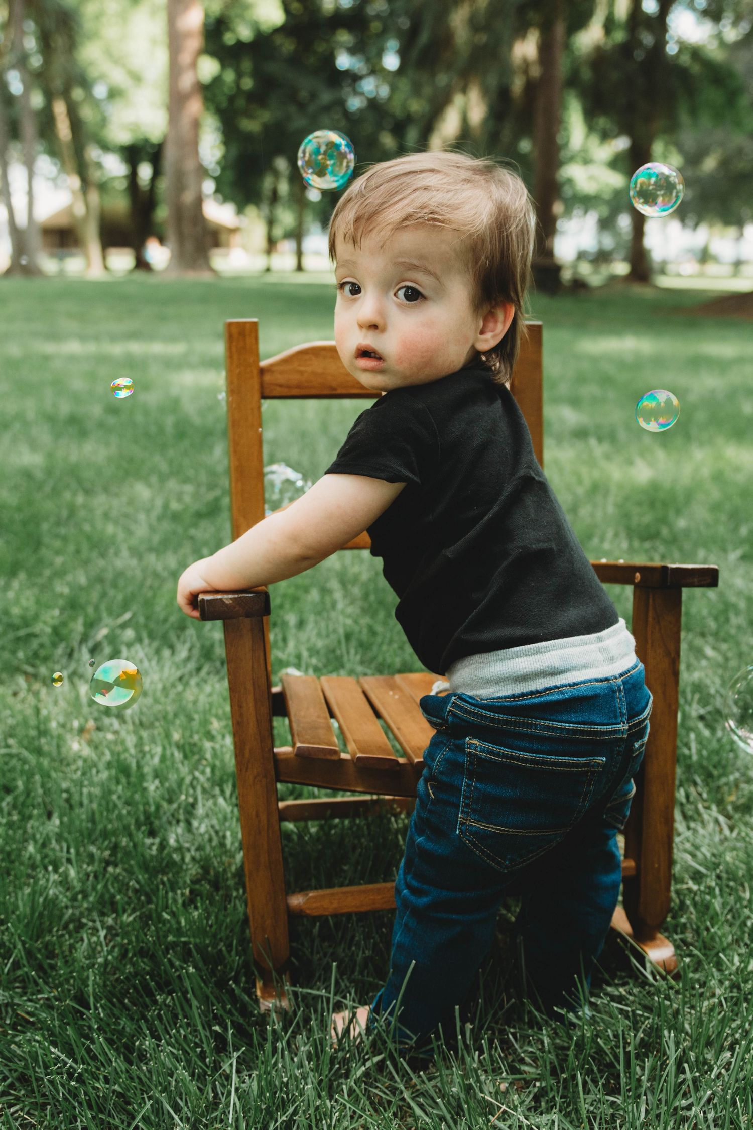 A toddler in denim and black stands by a wooden chair on grass while soap bubbles float in the sunlit garden.