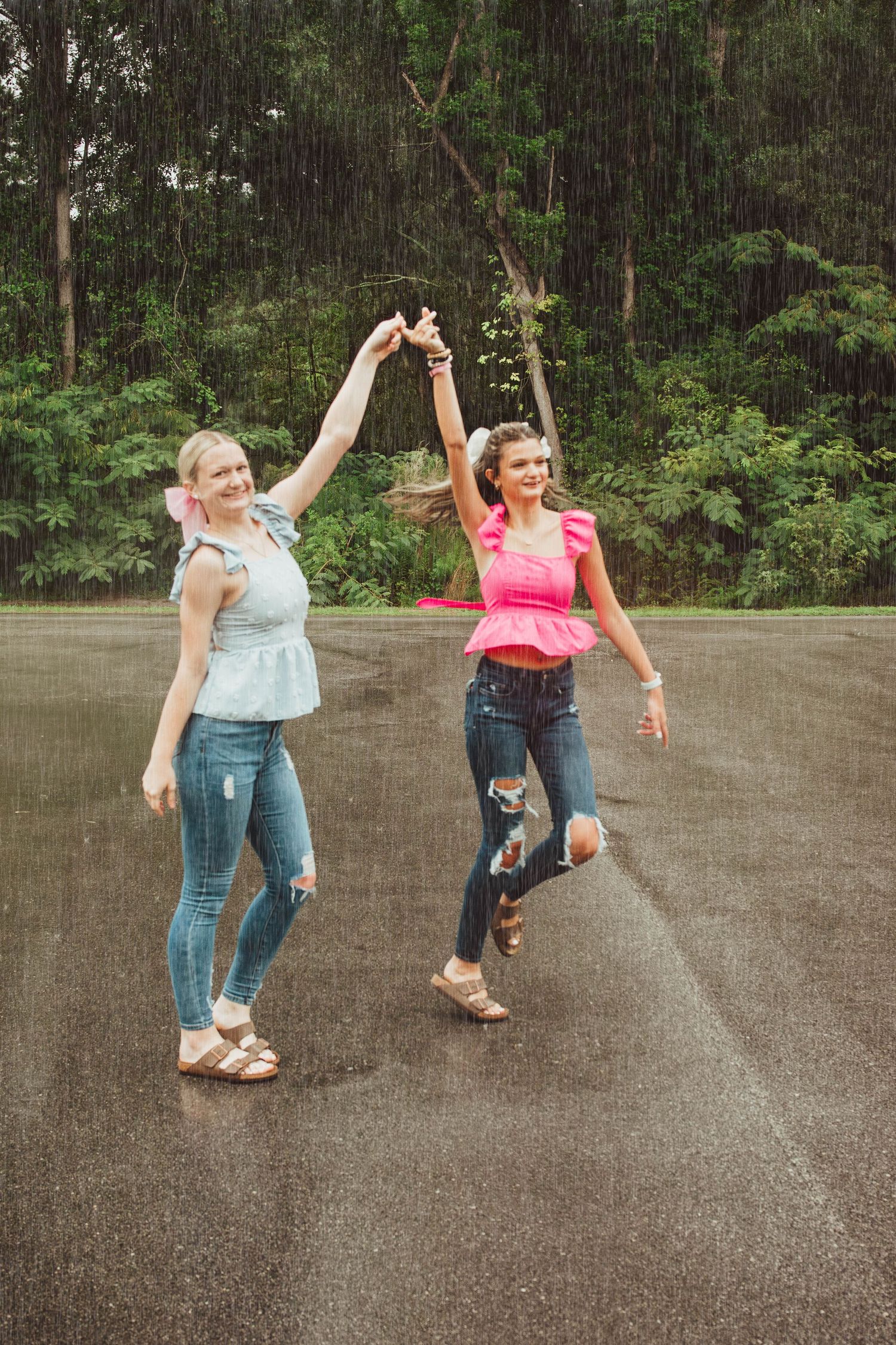 Two people dancing joyfully in casual outfits on a wet asphalt path with green forest in the background.