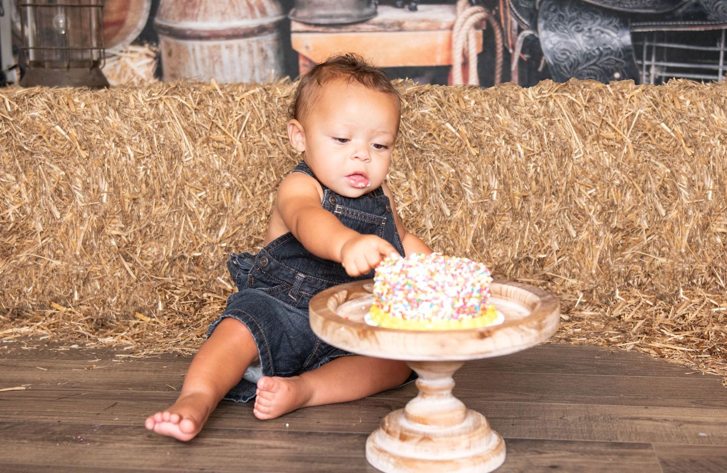 A young child in denim overalls sits on a wooden floor by hay bales reaching for a decorated cake on a pedestal stand.