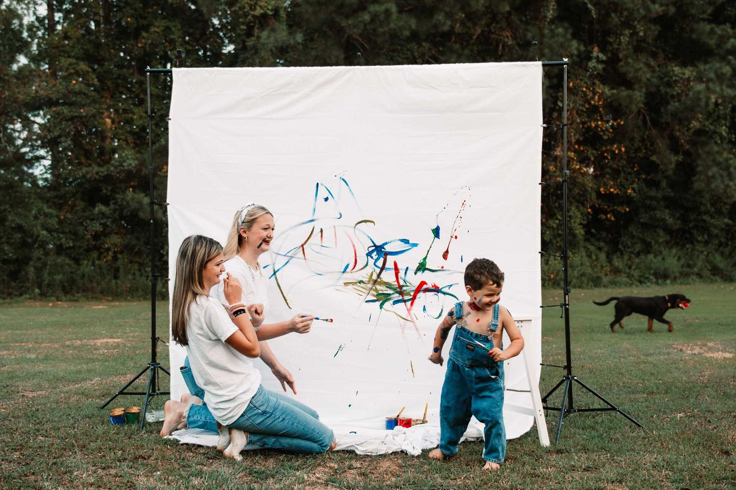 Small child in overalls creating colorful paint art on white canvas backdrop outdoors while being assisted.