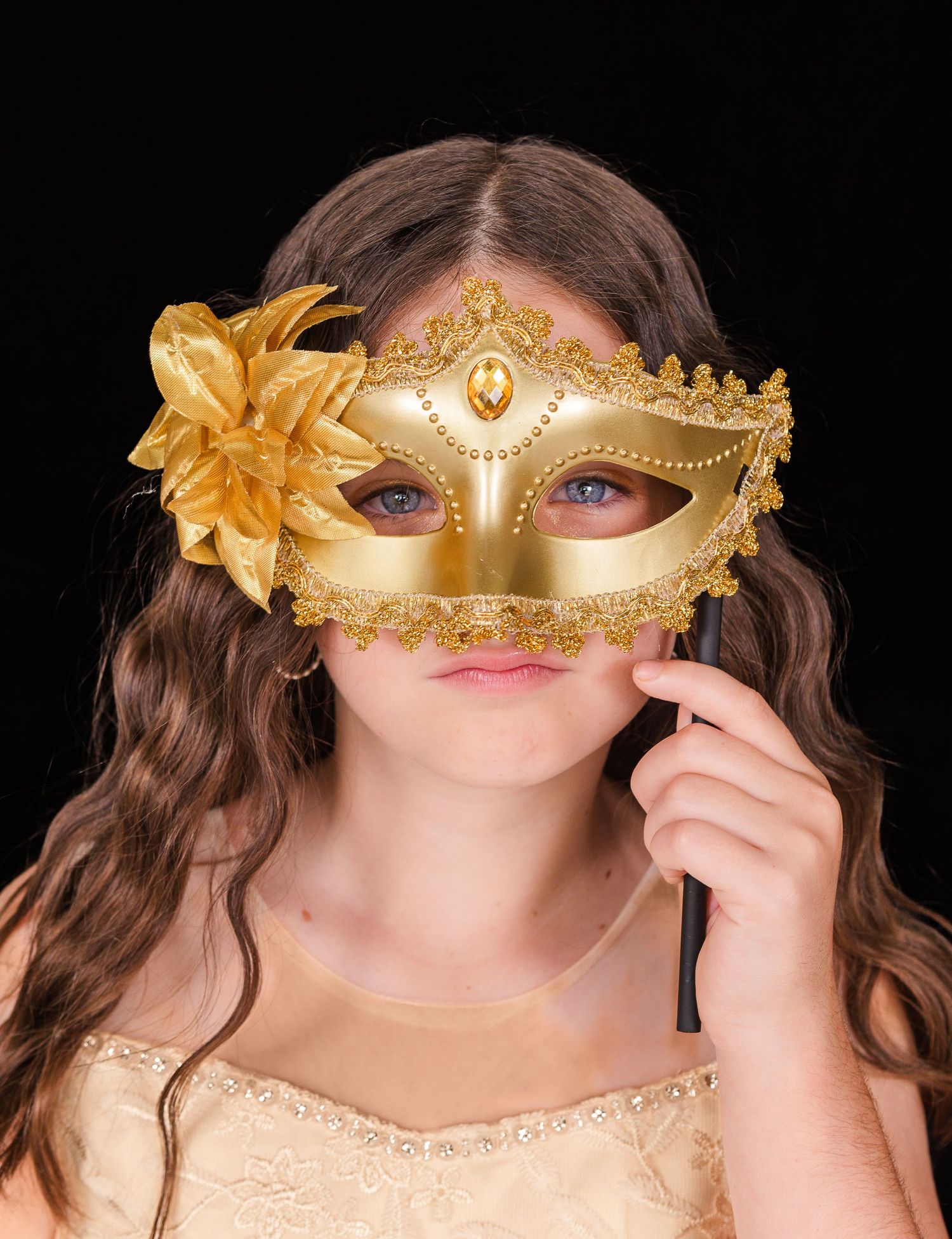 A person wearing a gold masquerade mask with floral decoration and glitter details poses in an elegant peach-colored dress.