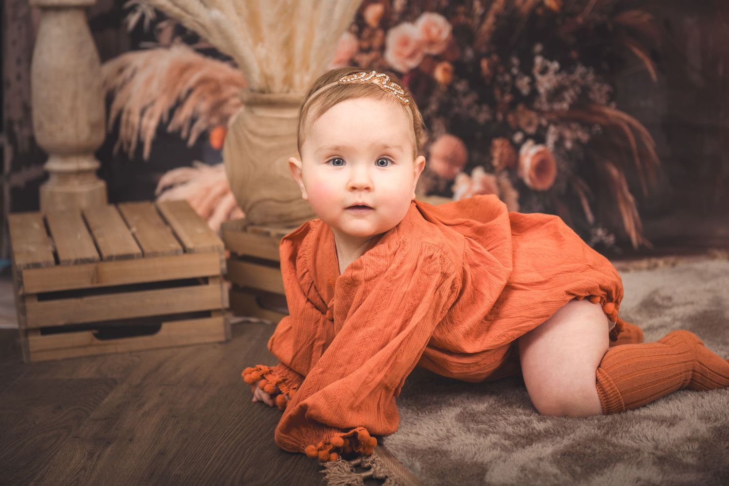 A baby in a ruffled orange romper crawls on a dark wooden floor against a rustic backdrop with flowers.