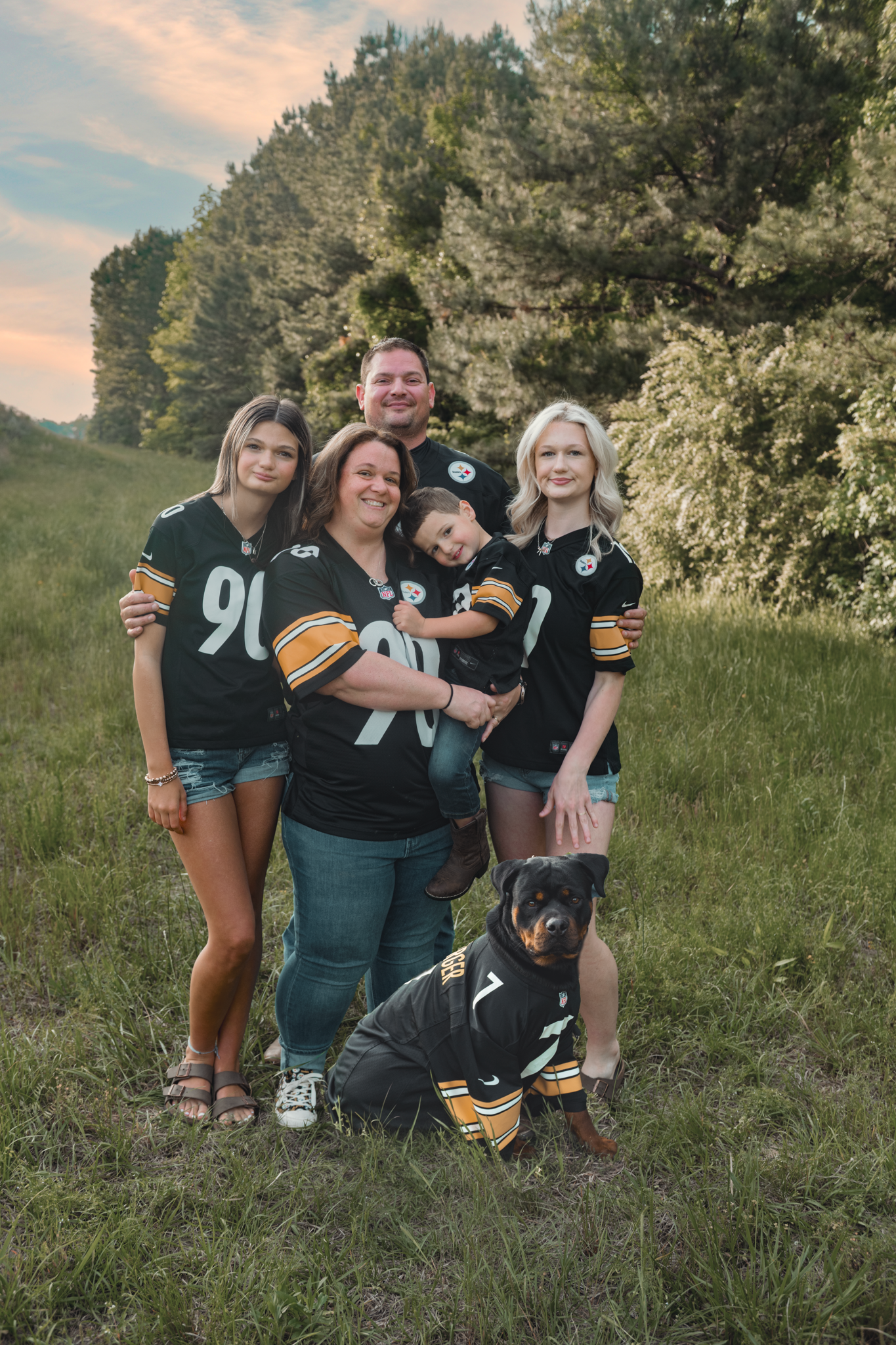 Outdoor group in matching Steelers or sports jerseys posing together with their black dog in a grassy field at dusk.