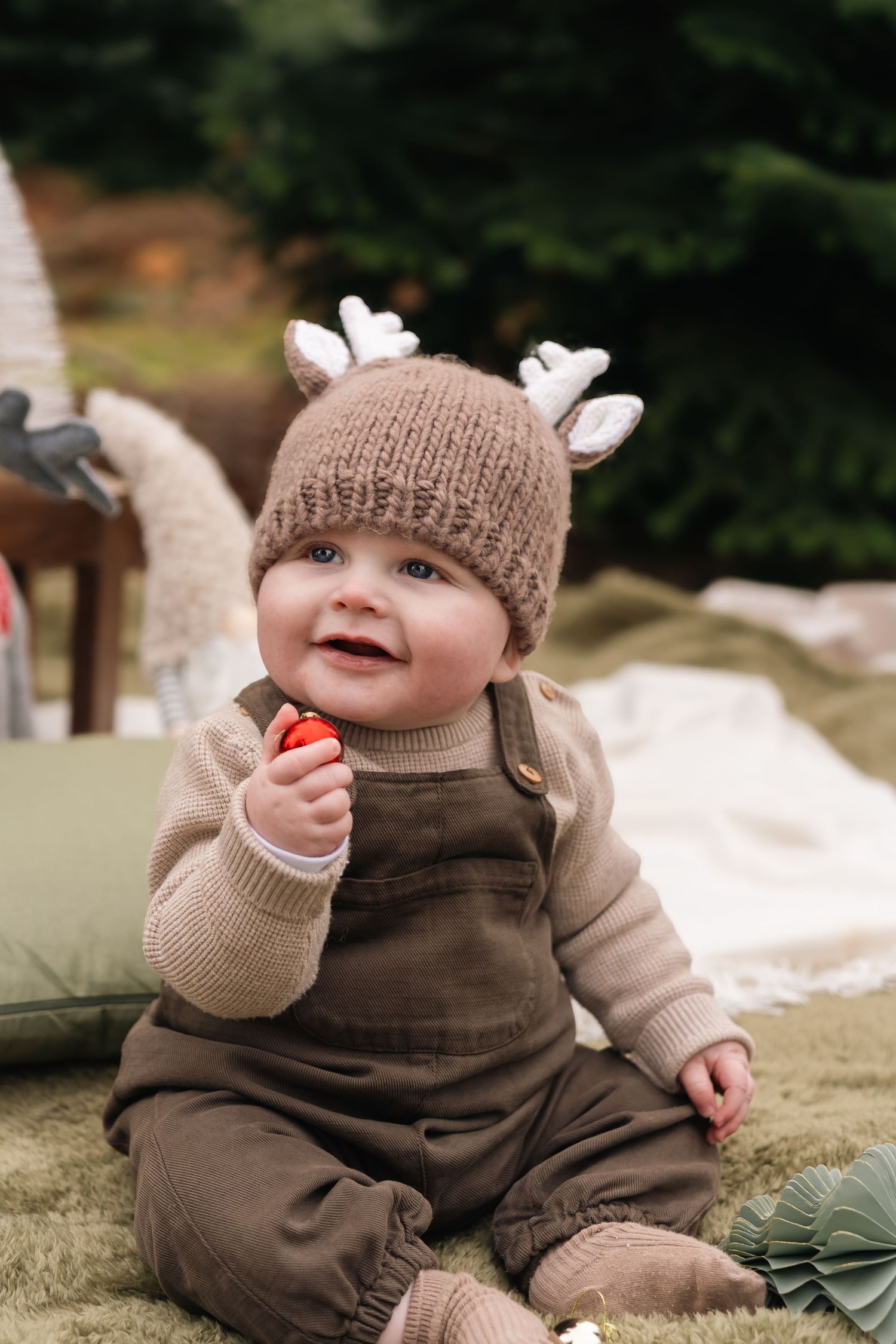 Adorable baby in brown reindeer knit hat and overalls sitting outdoors smiling during Christmas photo session.