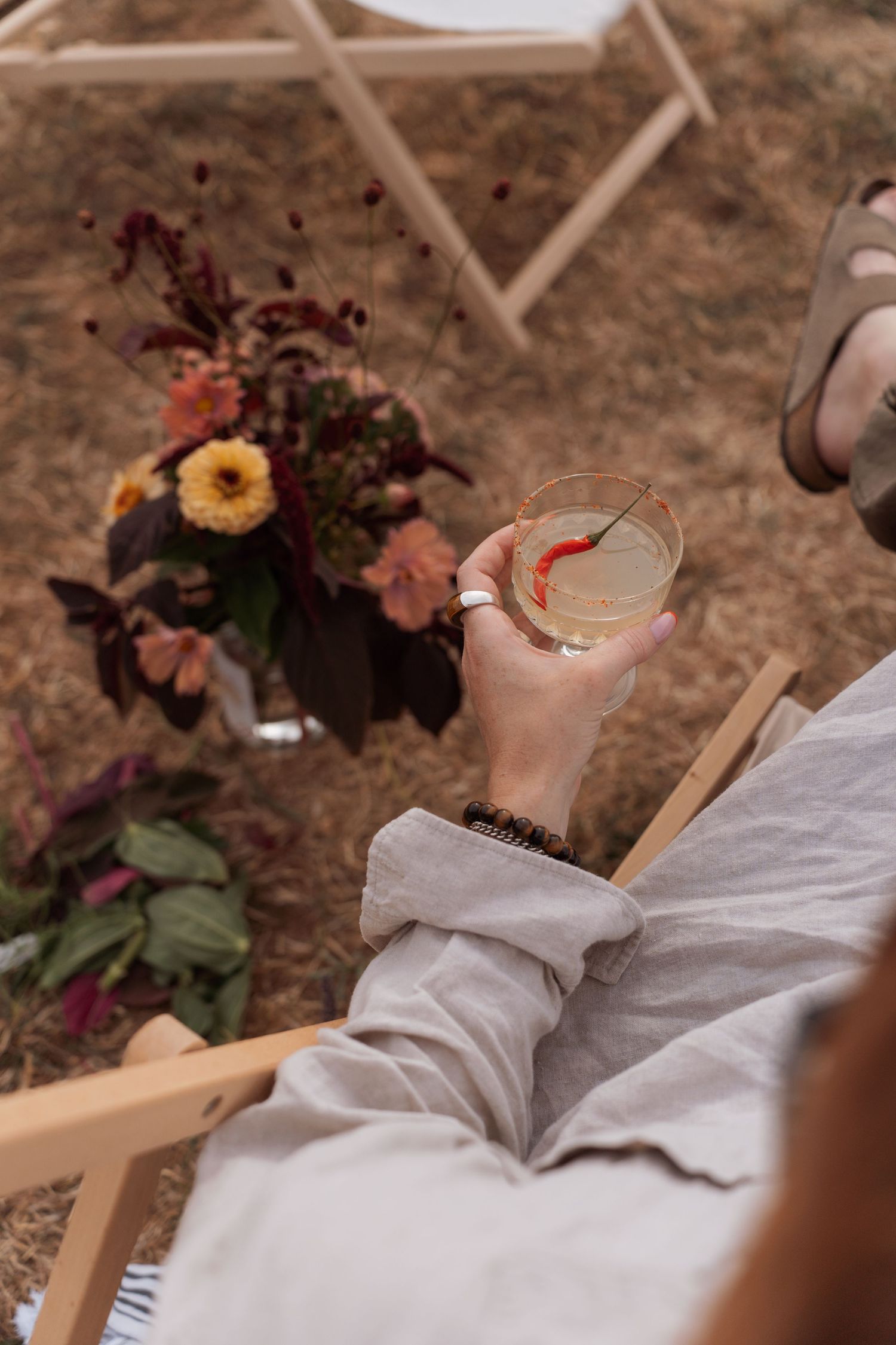 Person relaxing in wooden chair holding cocktail with chili pepper garnish near autumn floral arrangement outdoors.