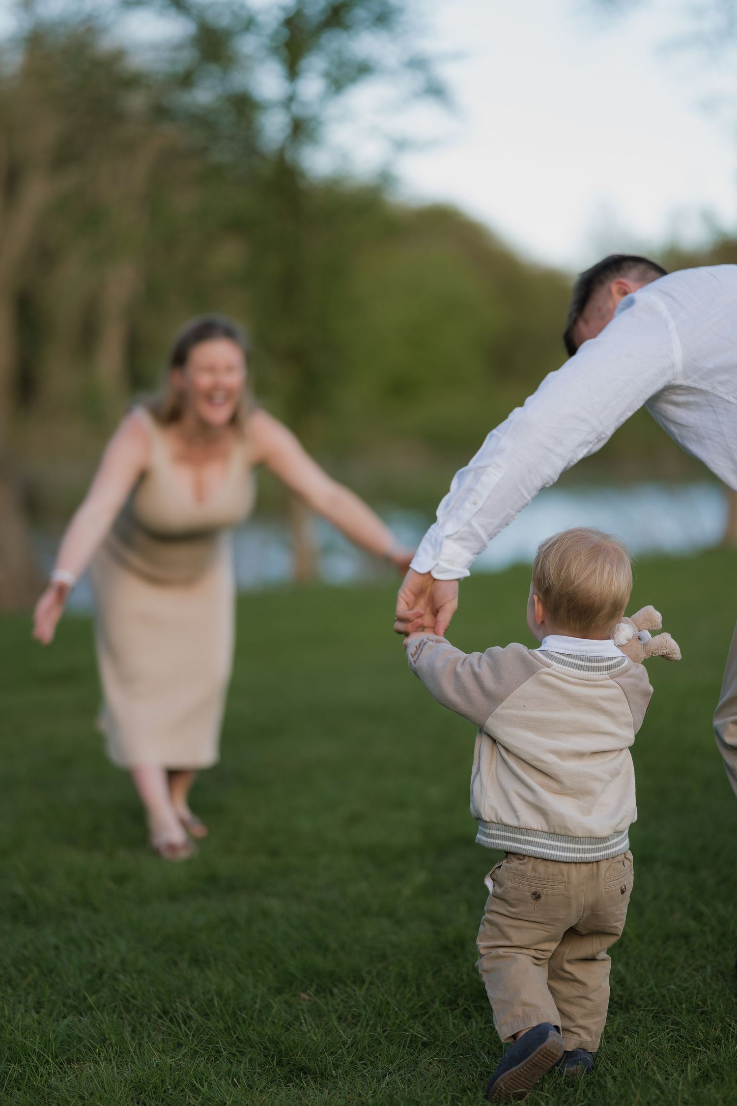A joyful family moment outdoors as a toddler walks toward laughing mom while dad holds his hand on green grass.