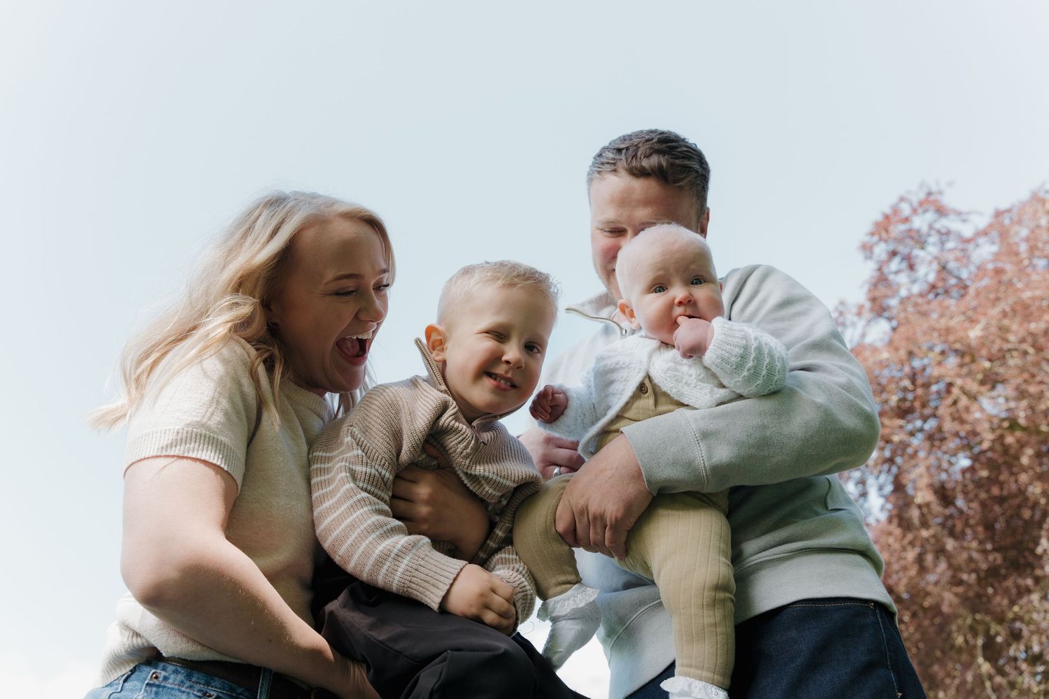 A joyful family with two young children laughing outdoors on a bright day, parents holding babies in casual clothing.
