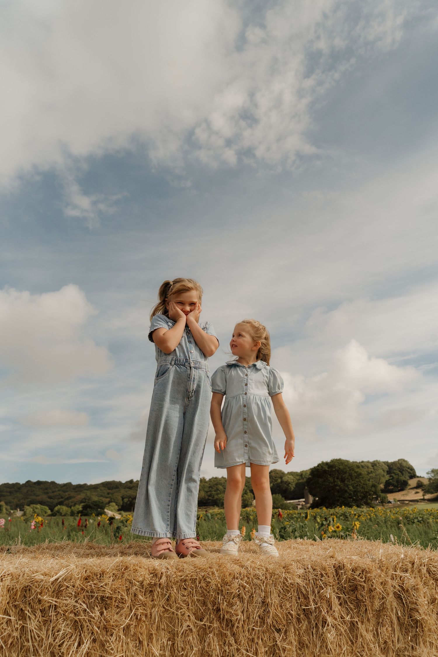 Two young girls in matching blue dresses stand on a hay bale in a sunny field with clouds and trees in the background.