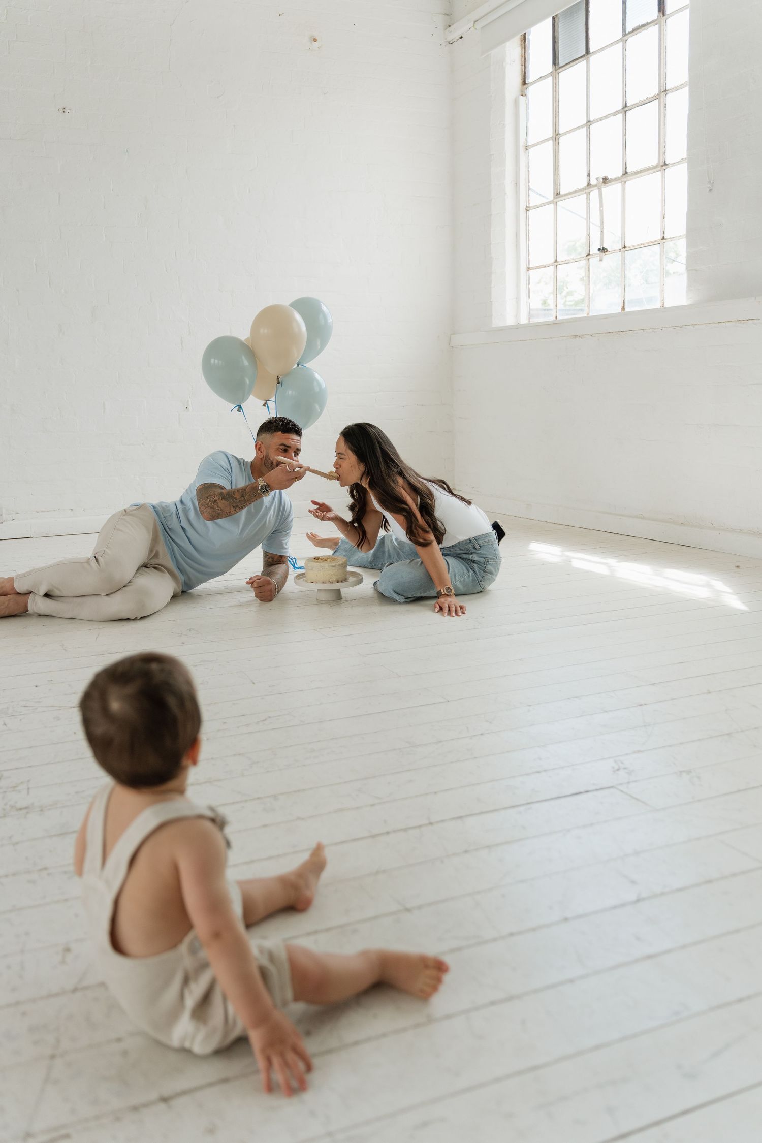 Family celebrating baby's first birthday on white floor with blue and beige balloons in bright studio.