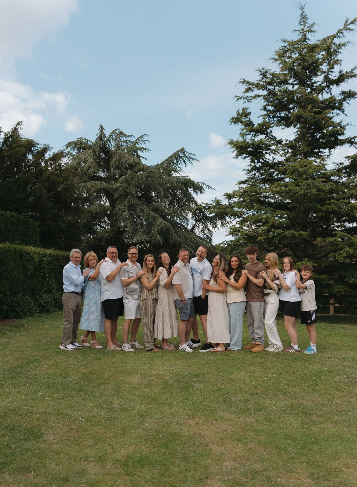 Large family group photo outdoors on green lawn with tall evergreen trees and blue sky background.