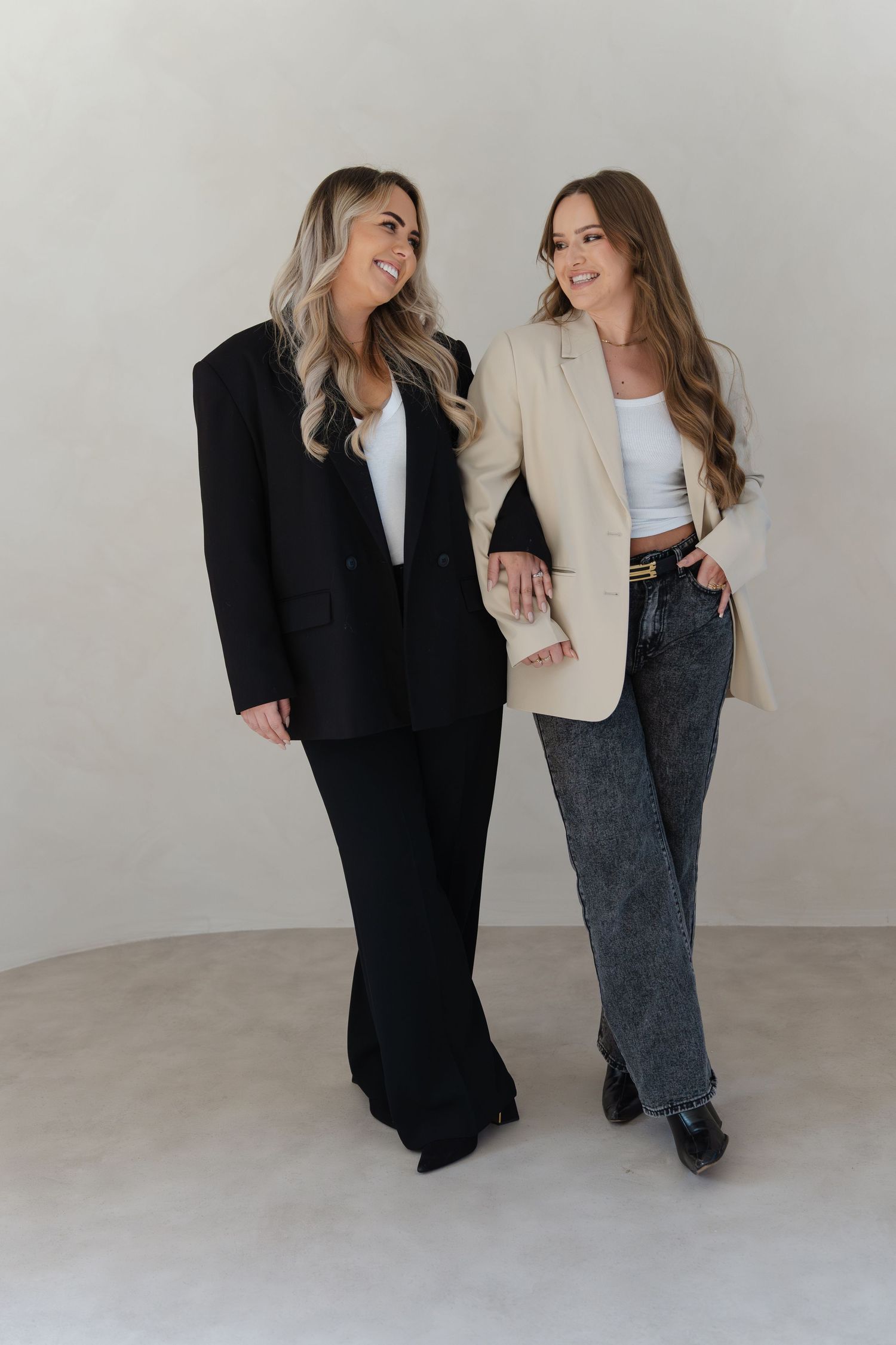 Two women in business attire laugh together while posing against a light gray studio backdrop.
