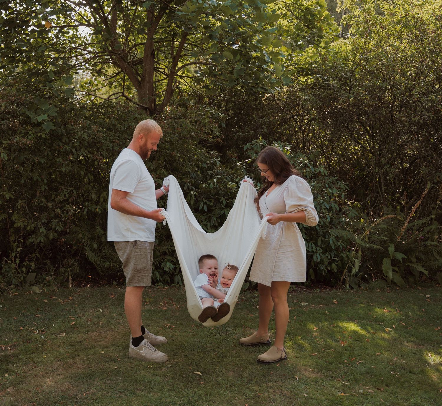 Couple holding a large white fabric with a baby photo print outdoors in a lush green garden setting.