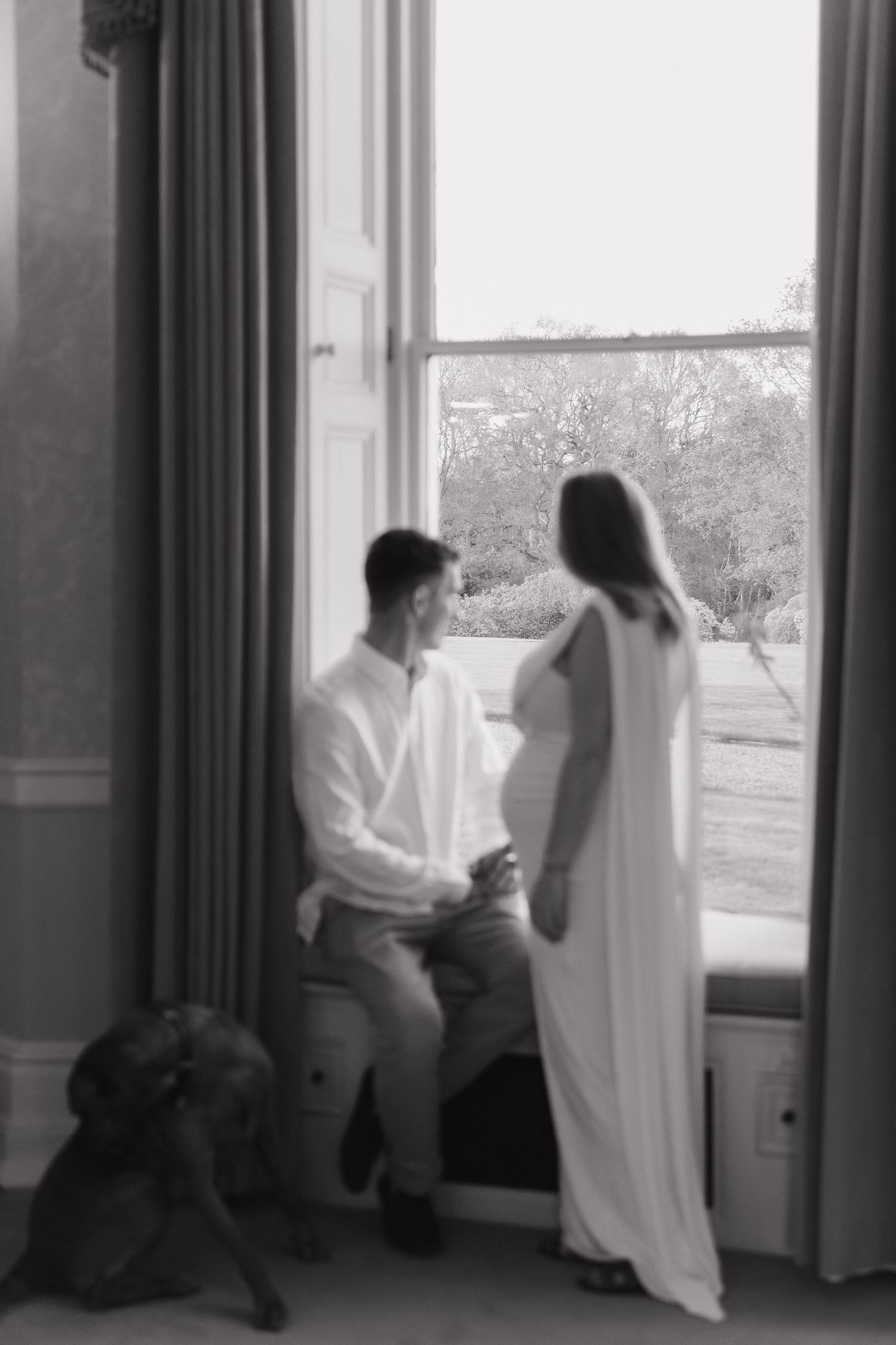Black and white photo of a couple by a large window, woman in white dress with baby bump, man seated, dog nearby.