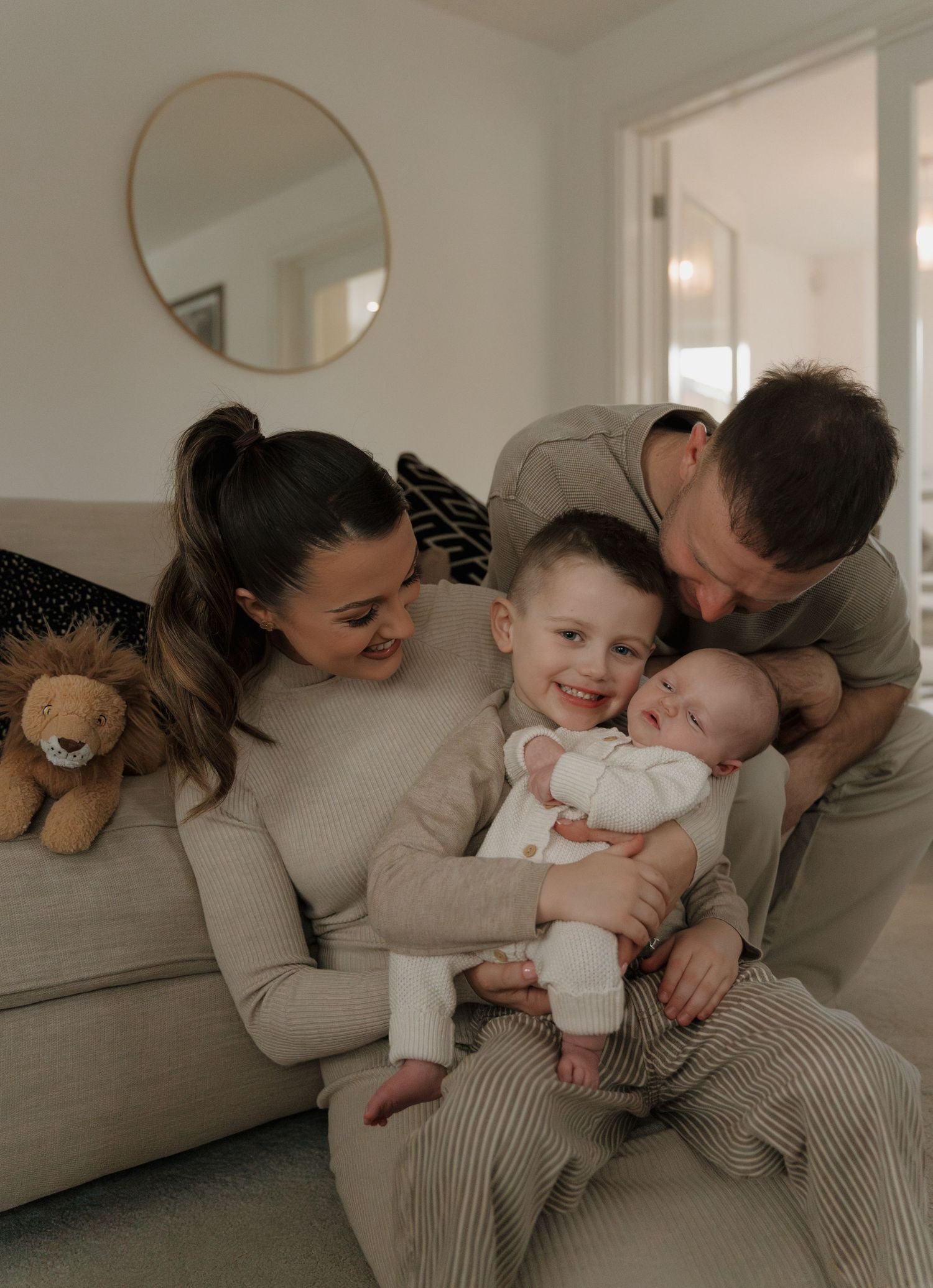 A happy family of four sits on a couch, with parents and toddler cuddling a newborn baby in a cozy home.
