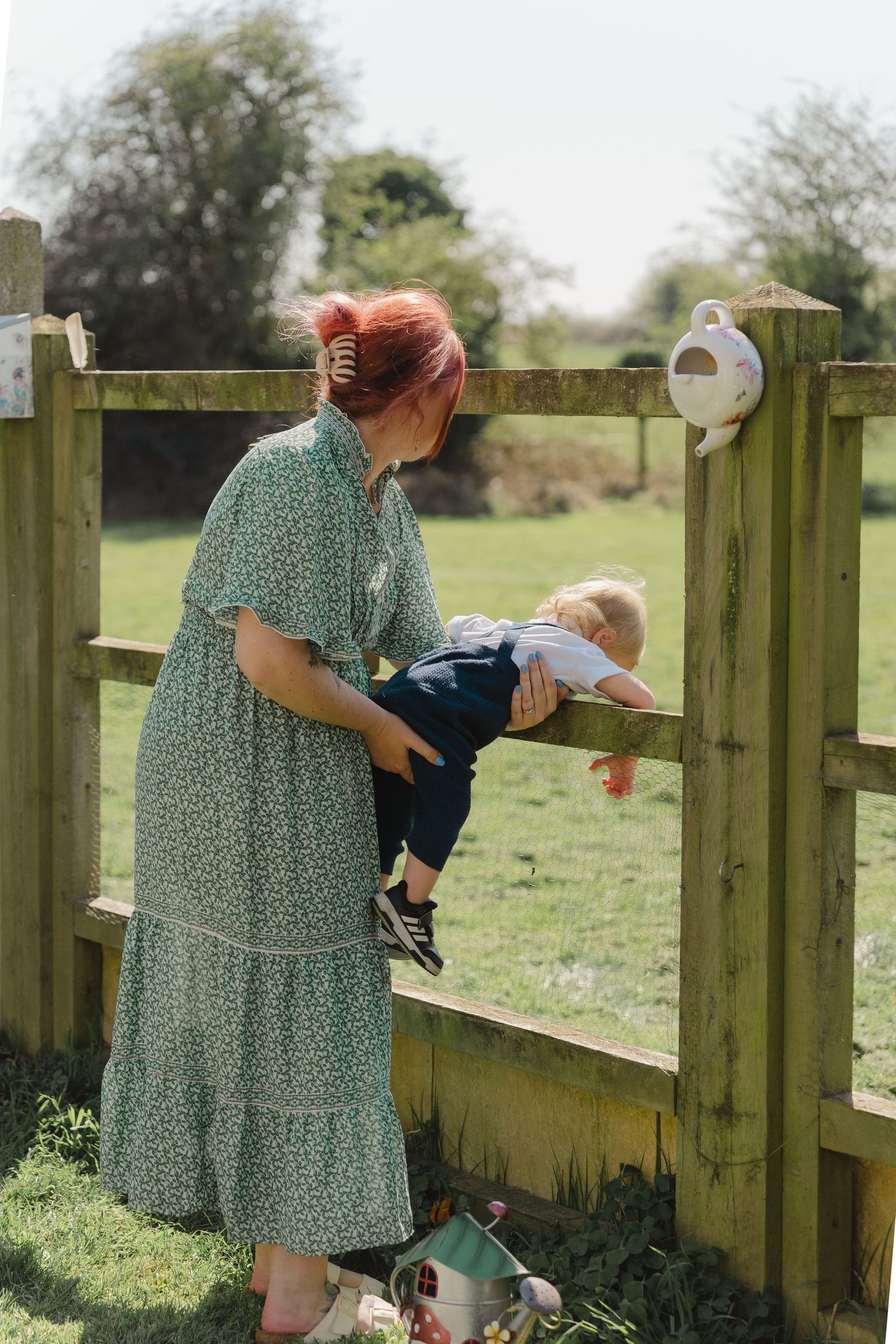 A person in a green floral dress stands at a wooden fence in a sunny meadow while holding a small child.