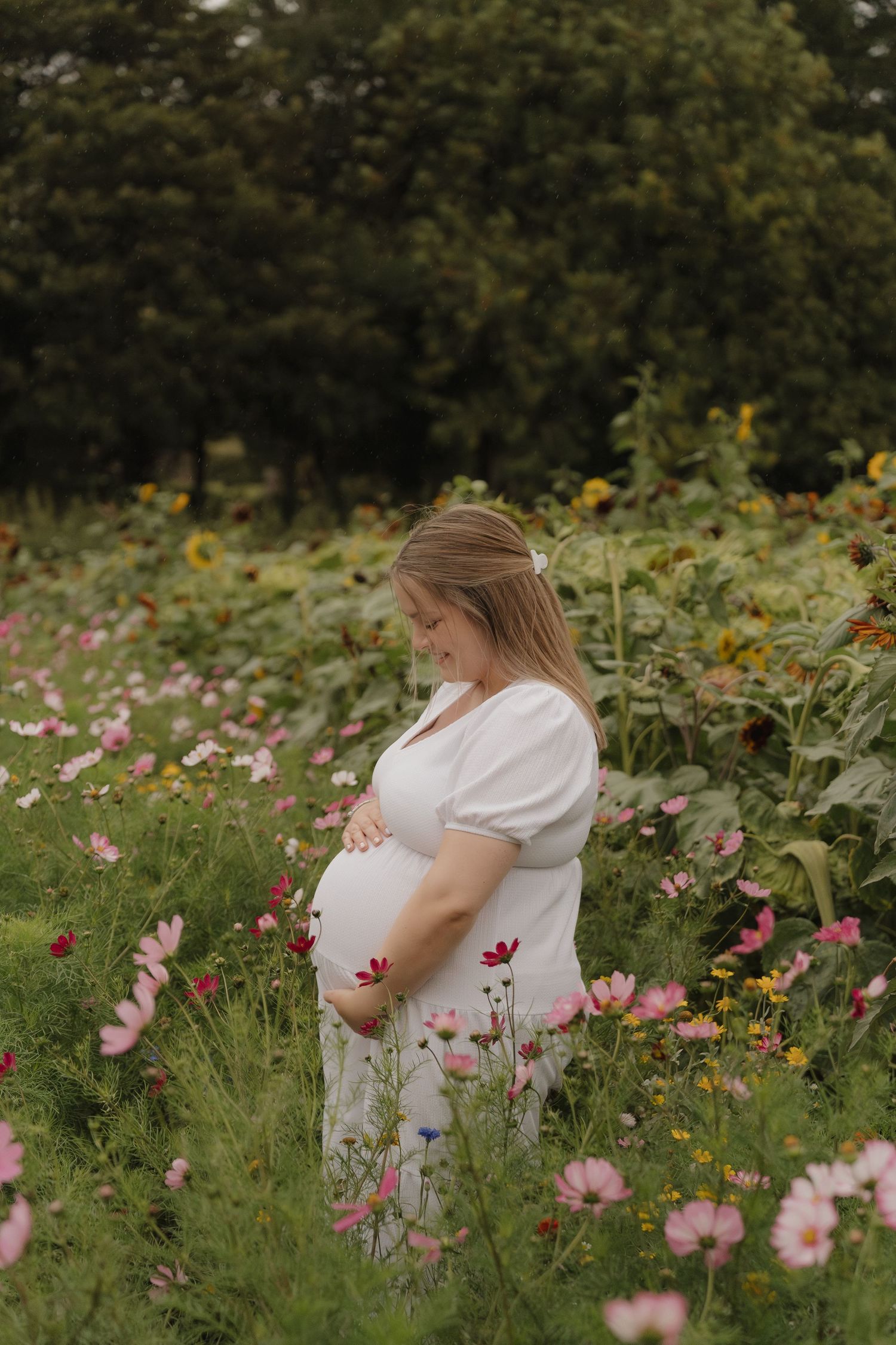 A pregnant woman in a white dress stands among pink cosmos flowers in a lush garden setting.