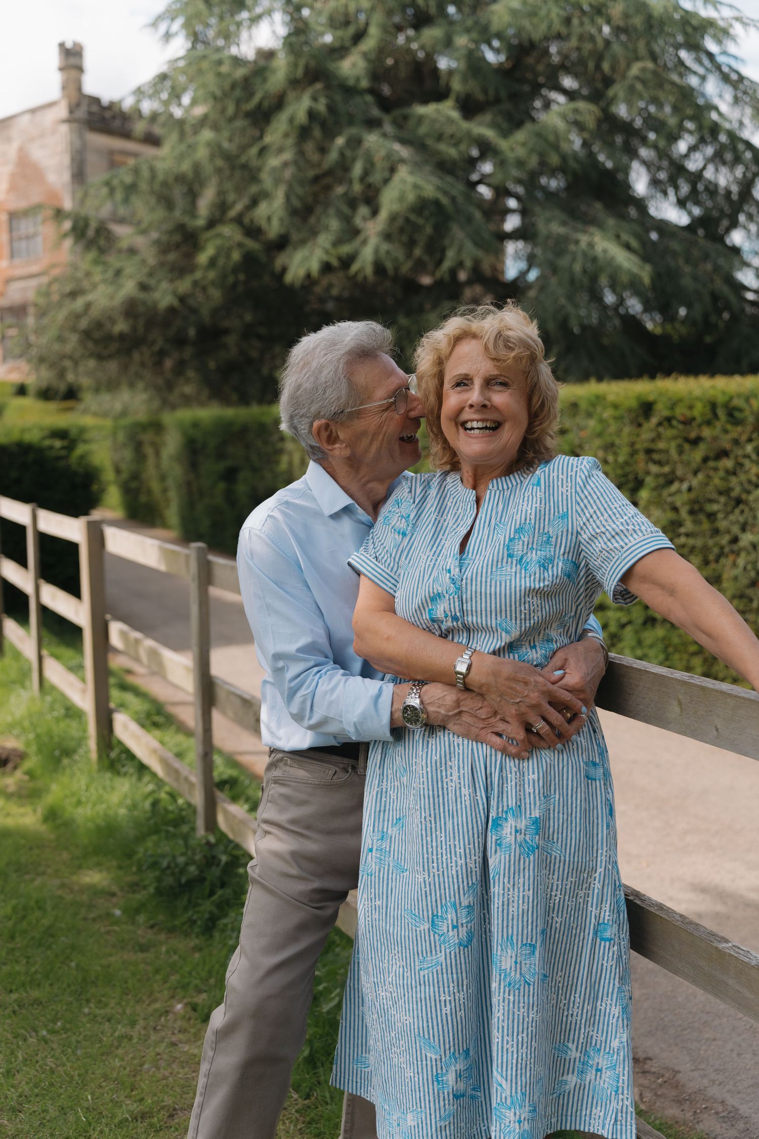 Elderly couple sharing a tender moment outdoors, man kissing woman's cheek as she smiles, standing by a wooden fence.
