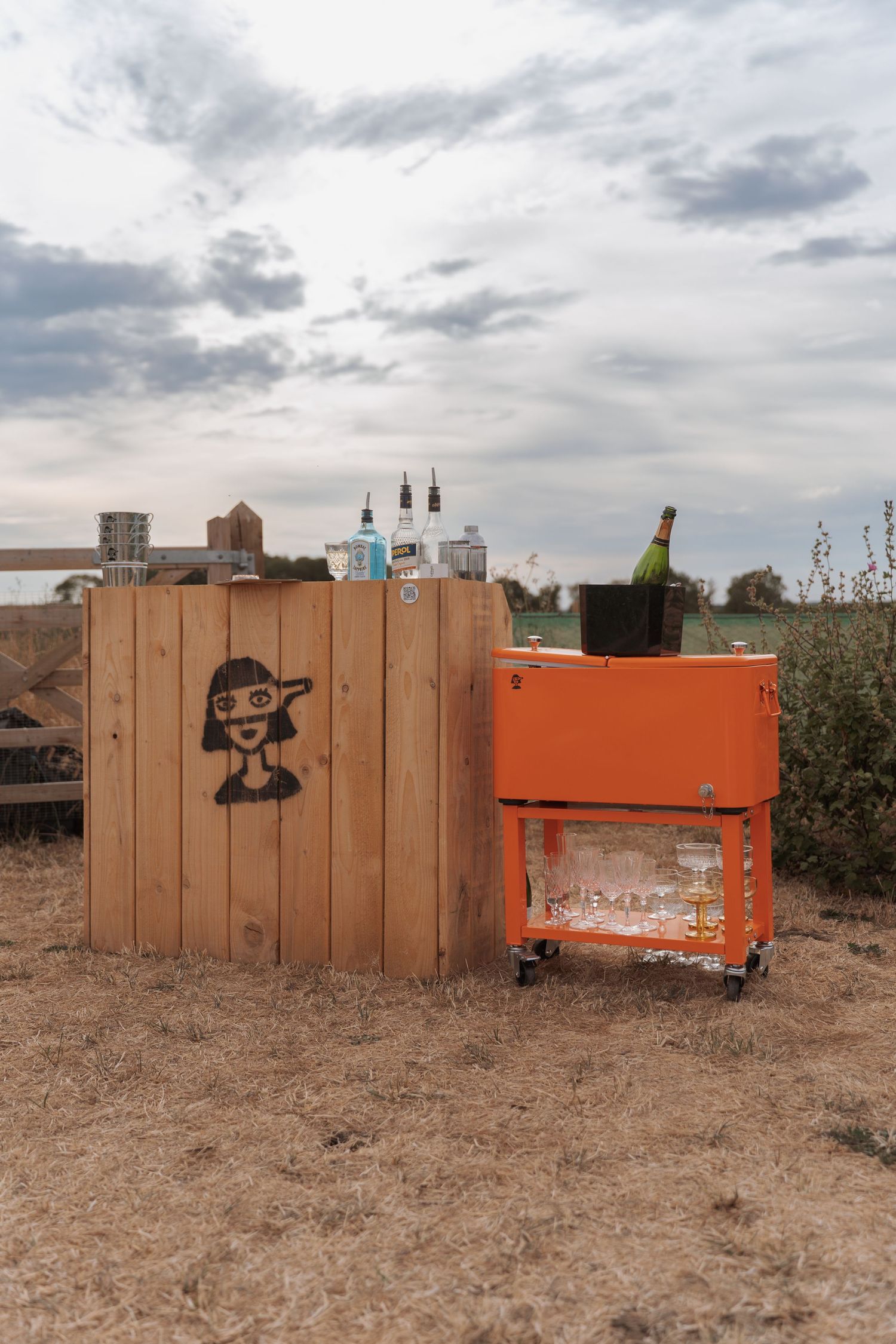 Outdoor bar setup with wooden pallet backdrop, cartoon graffiti, liquor bottles, and orange cart with champagne on ice.